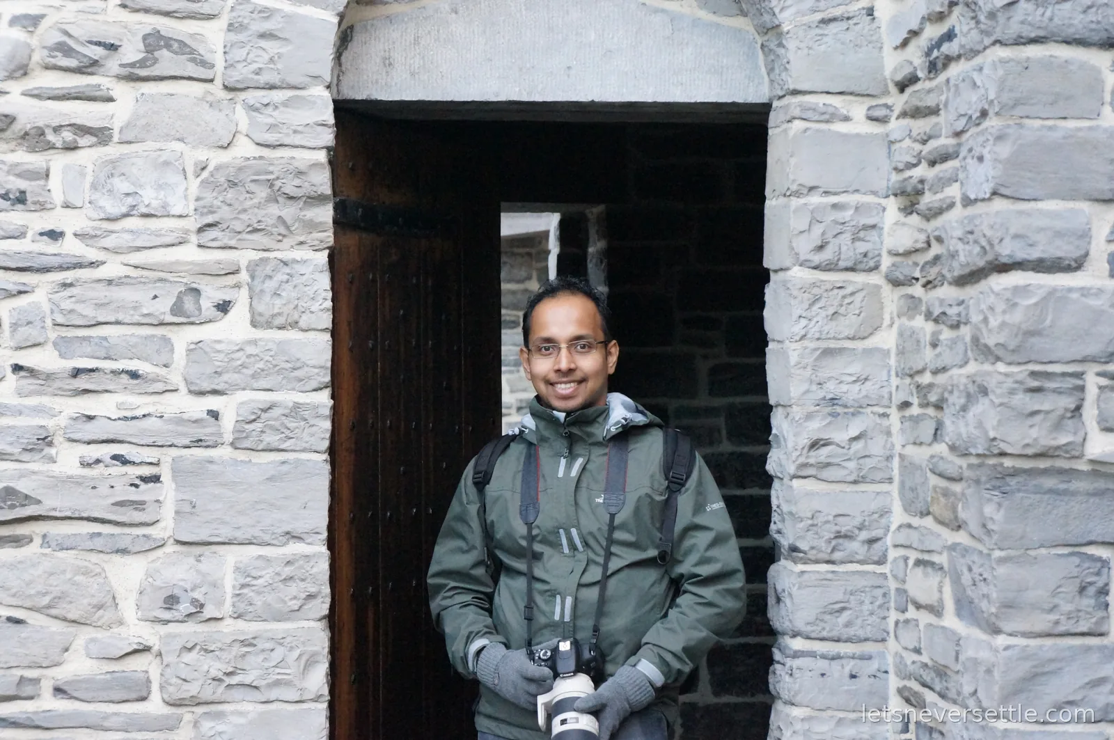 Smiling man in a green rain jacket and grey gloves holding a DSLR camera in an arched stone doorway at Gravensteen castle