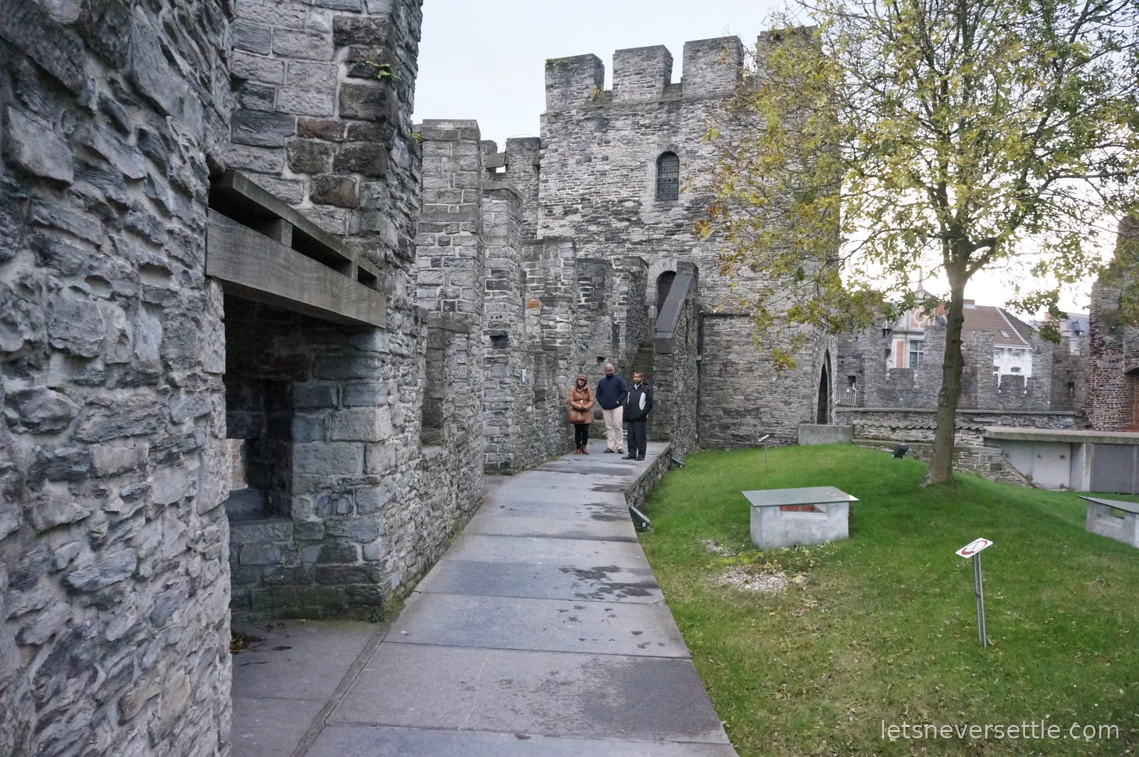 Battlements and wall-walk inside Gravensteen castle in Ghent, with three visitors on the stone path beside a small lawn