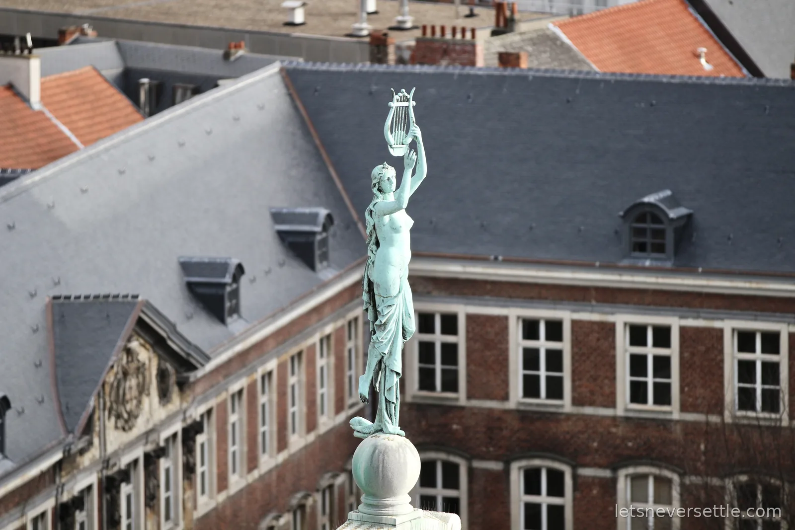 Weathered green statue of a muse holding a lyre overhead on a rooftop pedestal, brick facades and slate roofs of Ghent behind