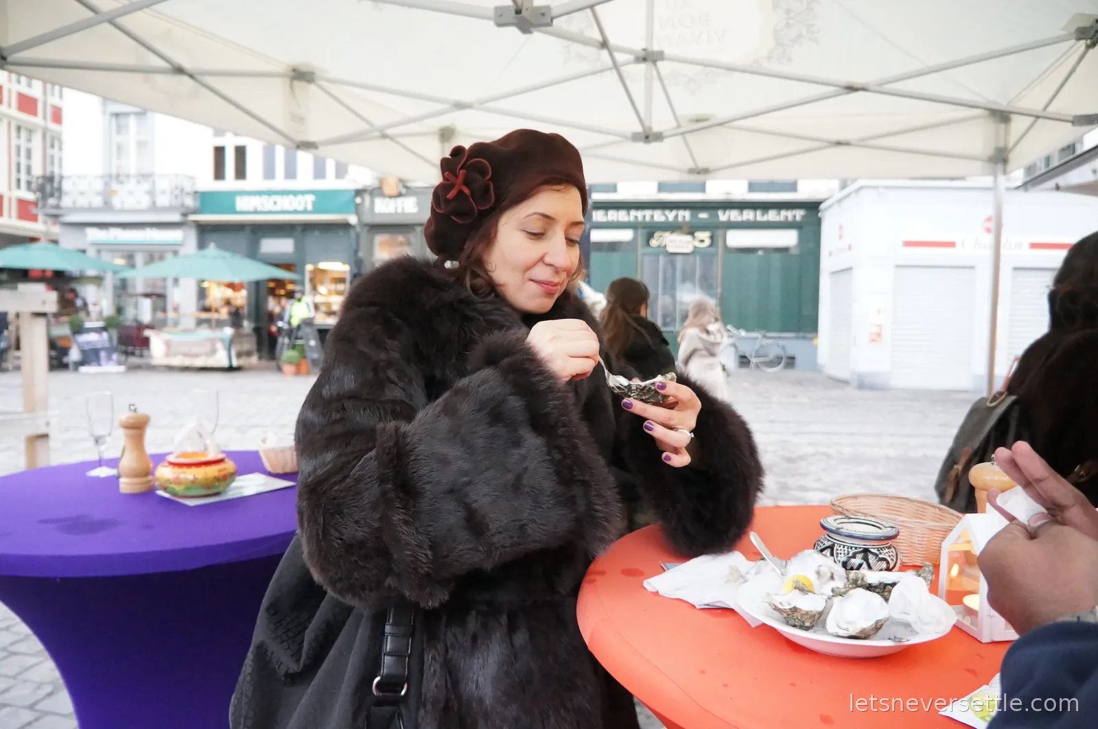 Inga in a dark fur coat and brown beret eating a fresh oyster at a standing table on the Groentenmarkt in Ghent, shopfronts behind