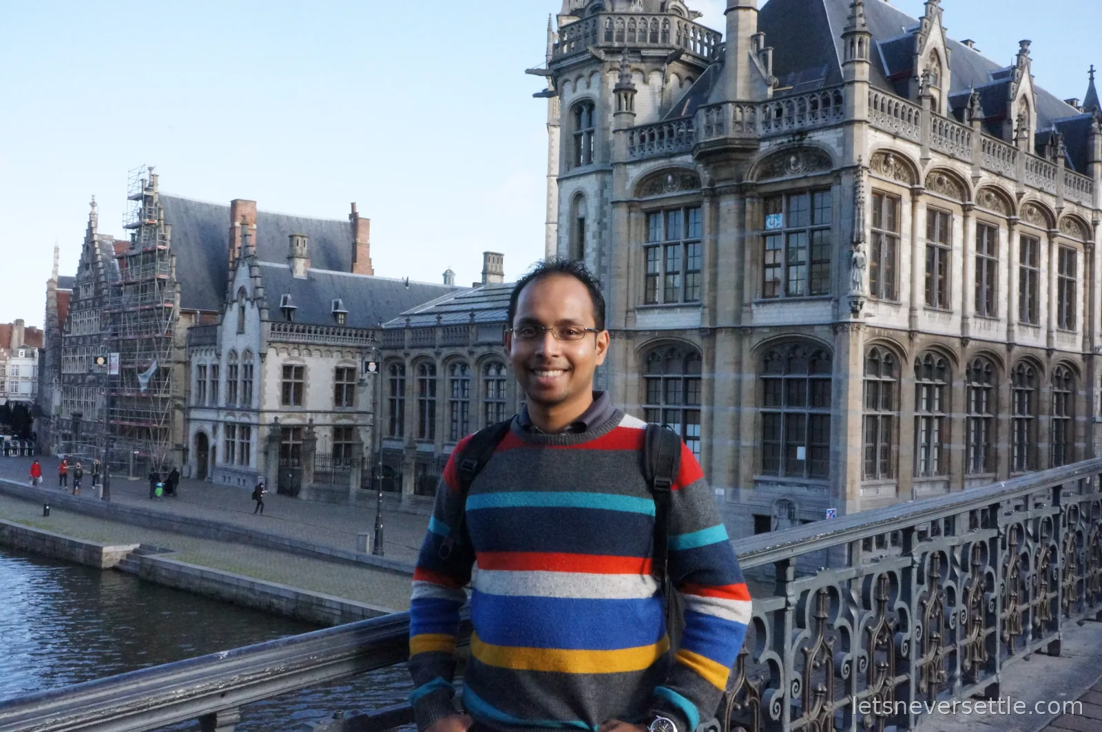 Man in a colourful striped jumper on St Michael's Bridge in Ghent, with the ornate stone old post office building and the canal behind