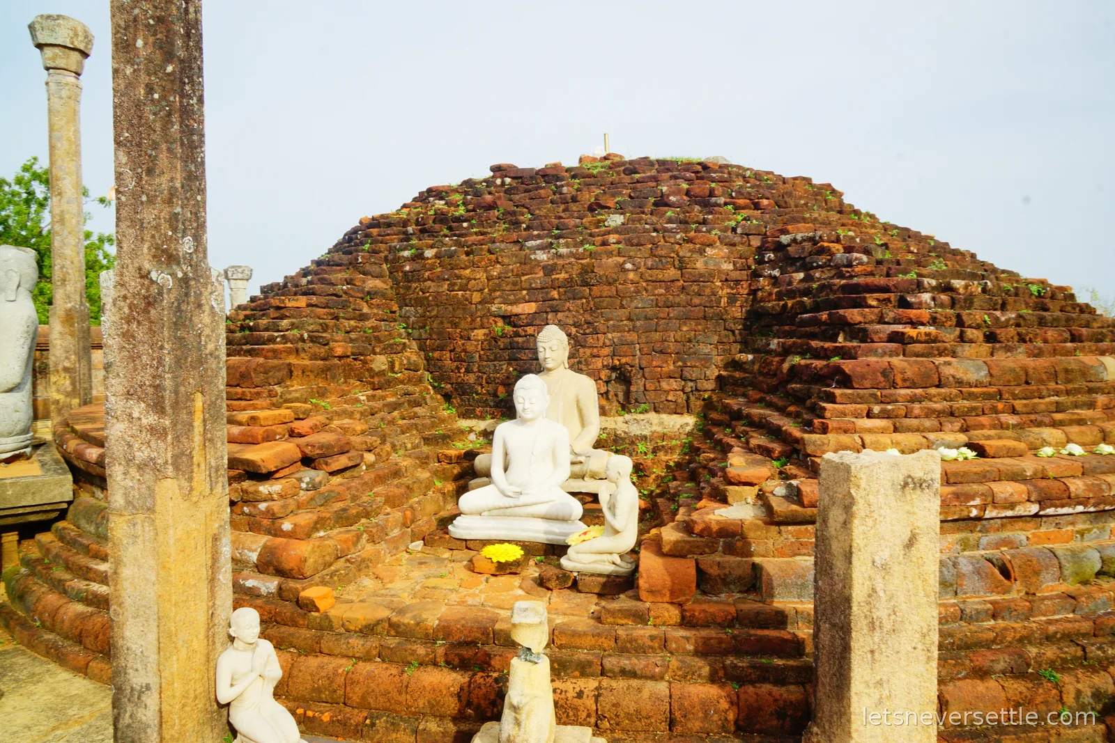 A ruined brick stupa at Girihandu Seya with its dome partly collapsed, small white Buddha and disciple statues placed on the brick steps in front