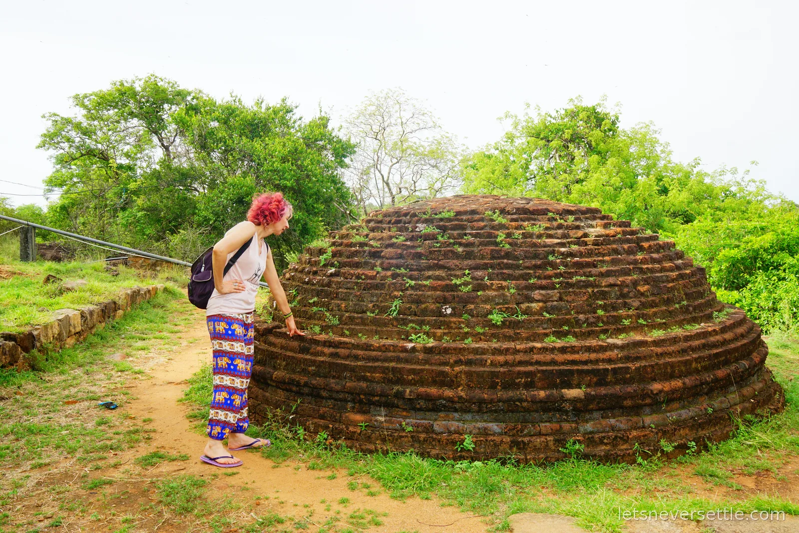 Inga in elephant-print trousers resting a hand on a small ruined brick stupa about her own height, grass and scrub behind it