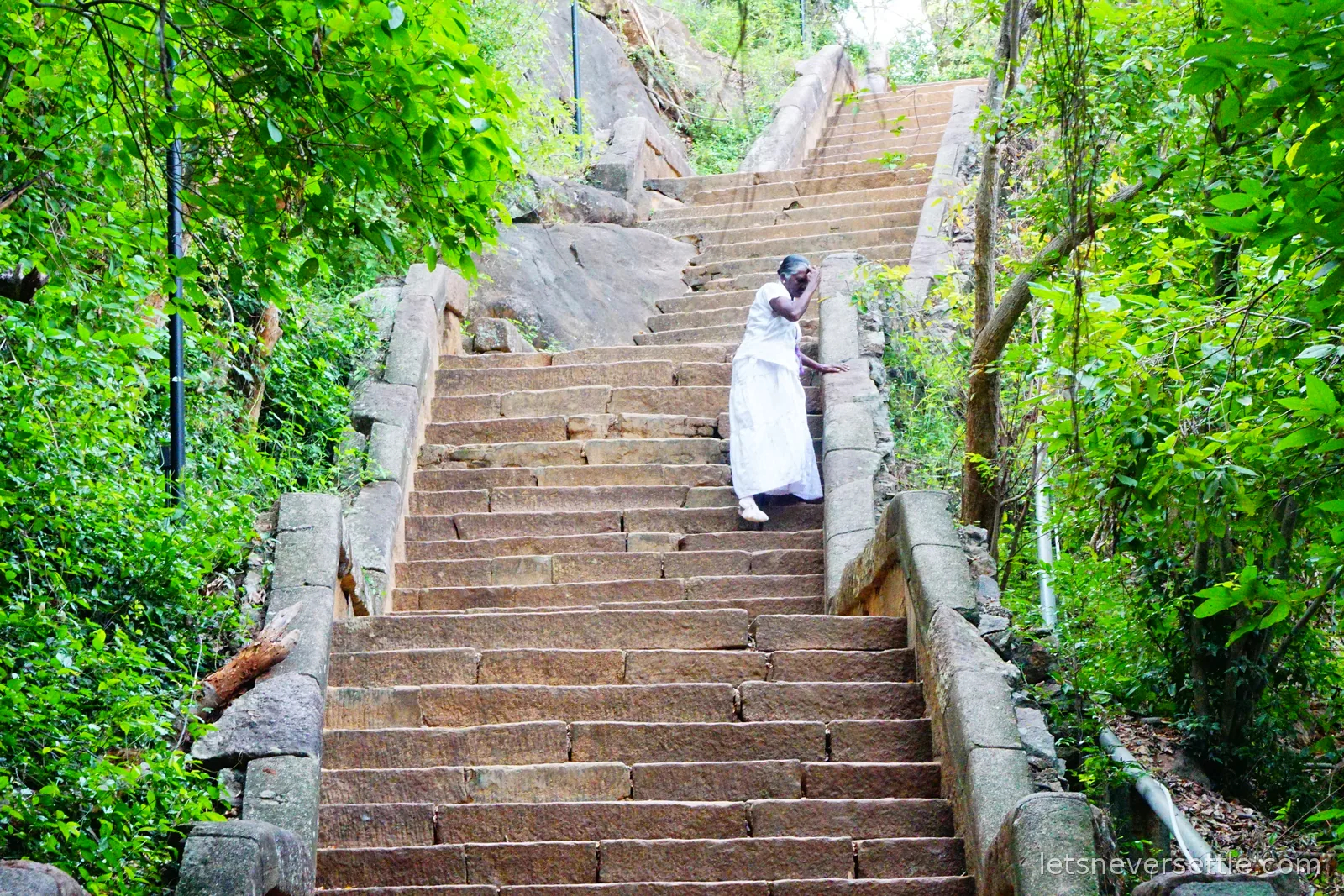 A steep flight of worn stone steps climbing between green trees at Girihandu Seya, with a woman in white walking down them