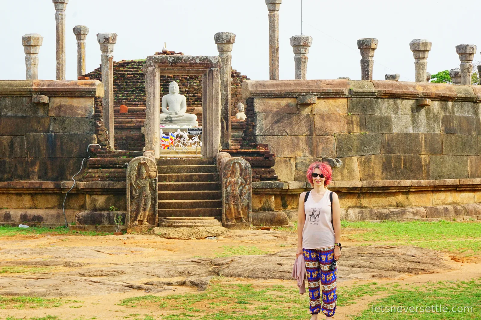 Inga standing on the bare rock in front of the vatadage, with a small white seated Buddha statue framed in the stone doorway behind her
