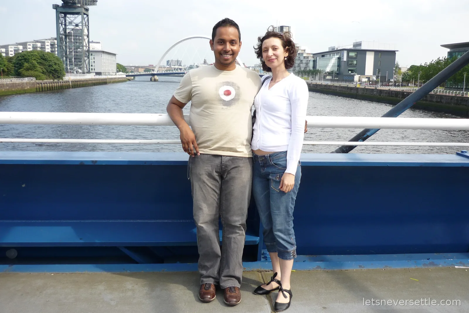 Inga and Dileep standing together on Bell's Bridge over the River Clyde, with the arched Clyde Arc bridge and the Finnieston Crane behind them