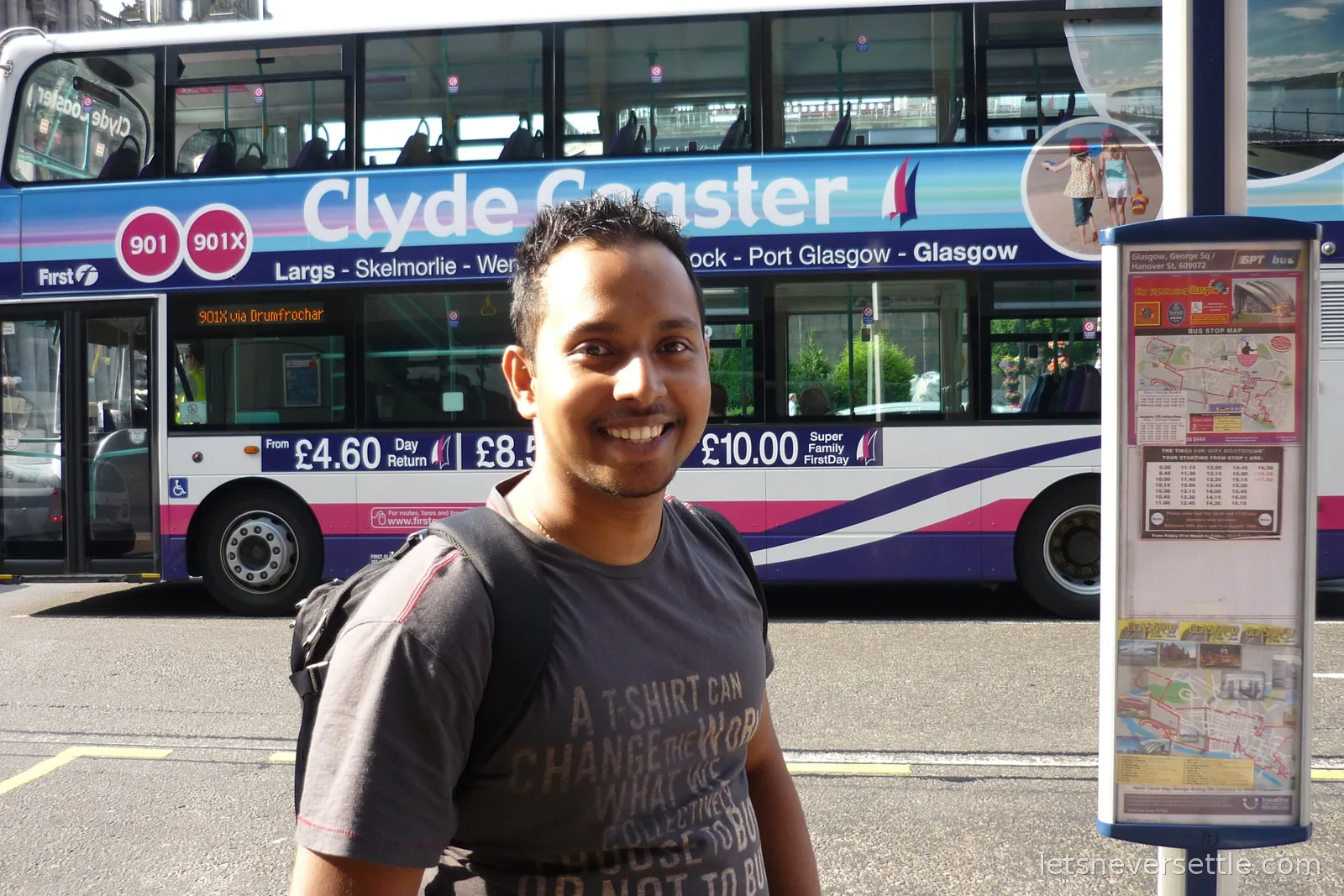 Dileep smiling at a George Square bus stop in Glasgow while a Clyde Coaster double-decker bus passes behind him
