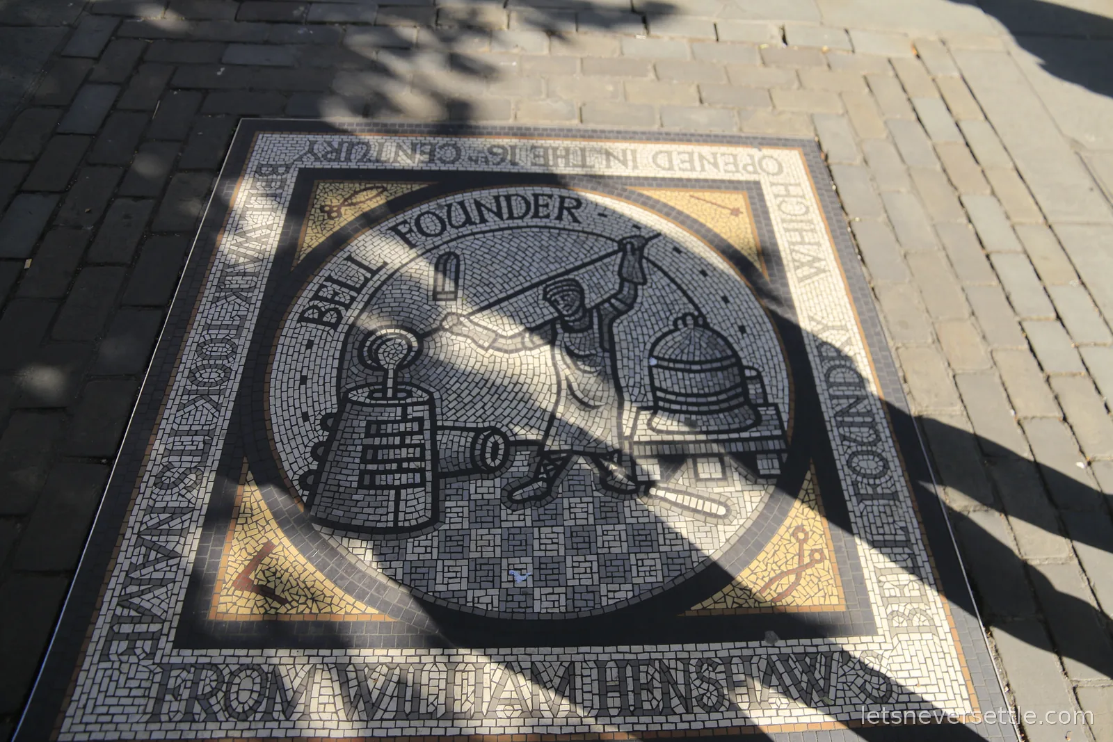 Pavement mosaic of a bell founder pouring molten metal beside a finished bell, with border lettering naming William Henshaw's bell foundry