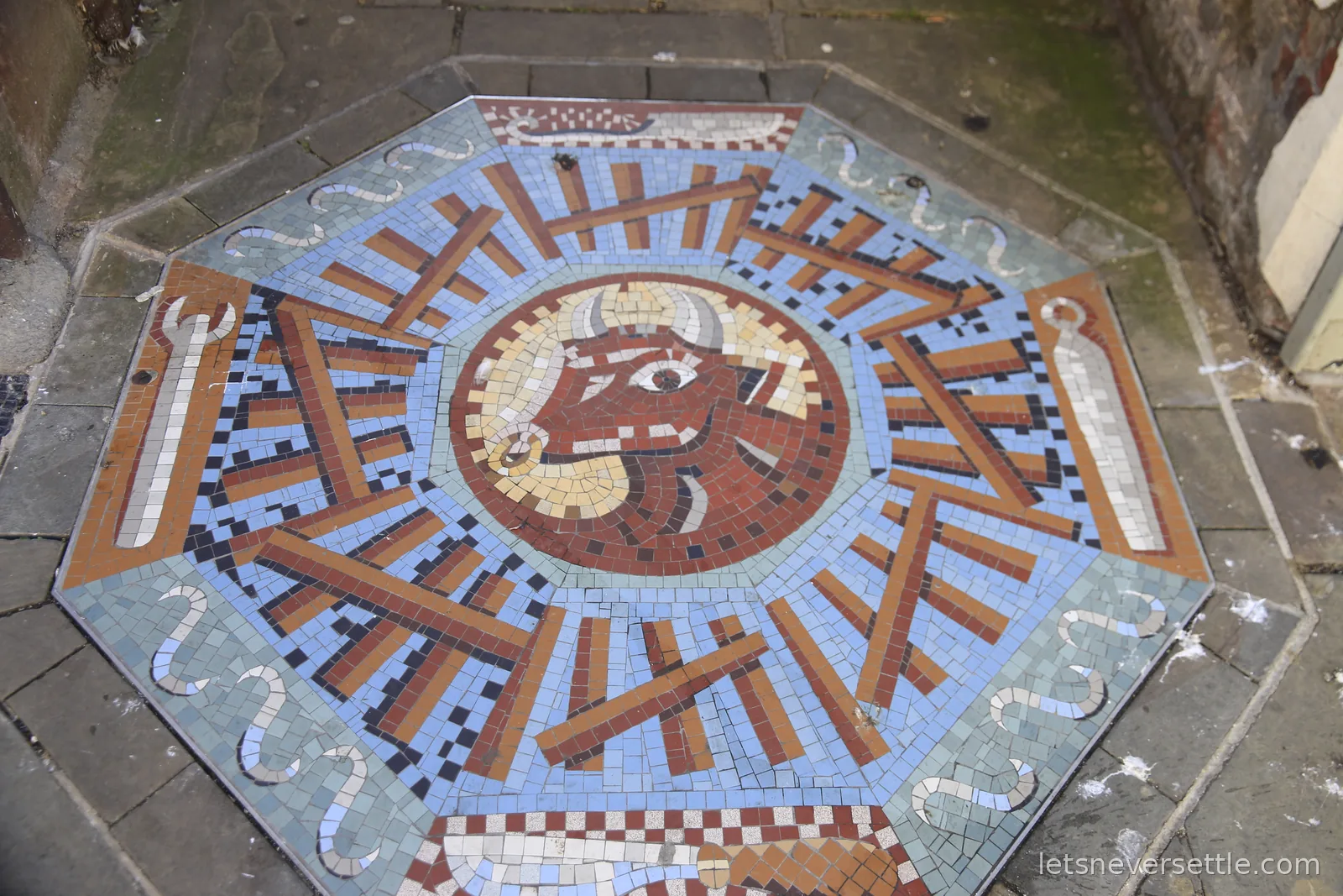 Octagonal pavement mosaic of a bull's head ringed by blue and terracotta tesserae, set into a stone pavement in Gloucester