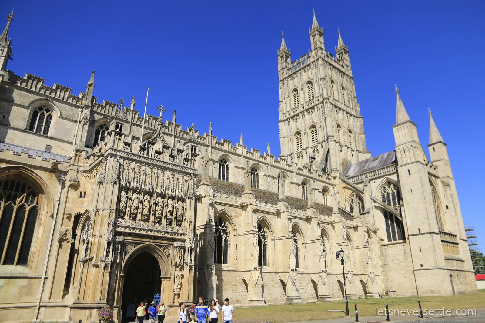 Gloucester Cathedral from the south, its carved entrance porch crowded with statues and the great pinnacled tower against a deep blue sky