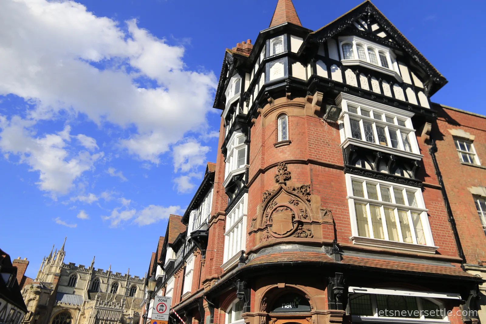Ornate red-brick corner building with black-and-white timbered upper storeys and a small spire in Gloucester city centre, the cathedral rising in the background
