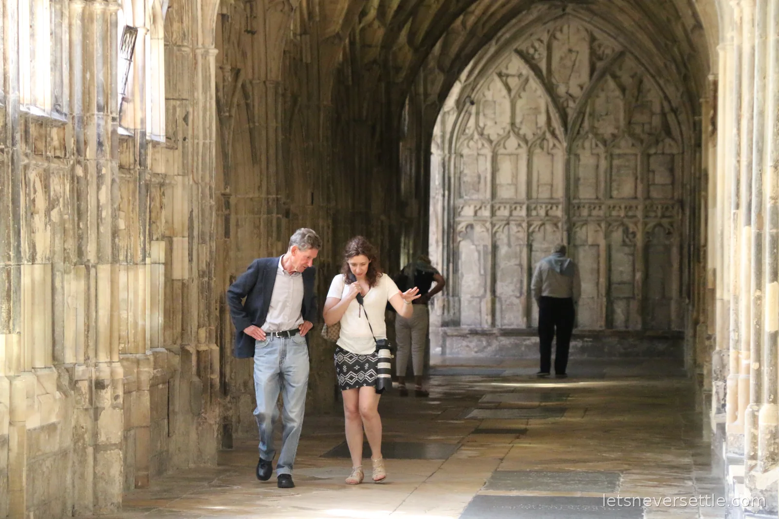 Visitors walking through the fan-vaulted cloister of Gloucester Cathedral, carved stone tracery panels lining the walk