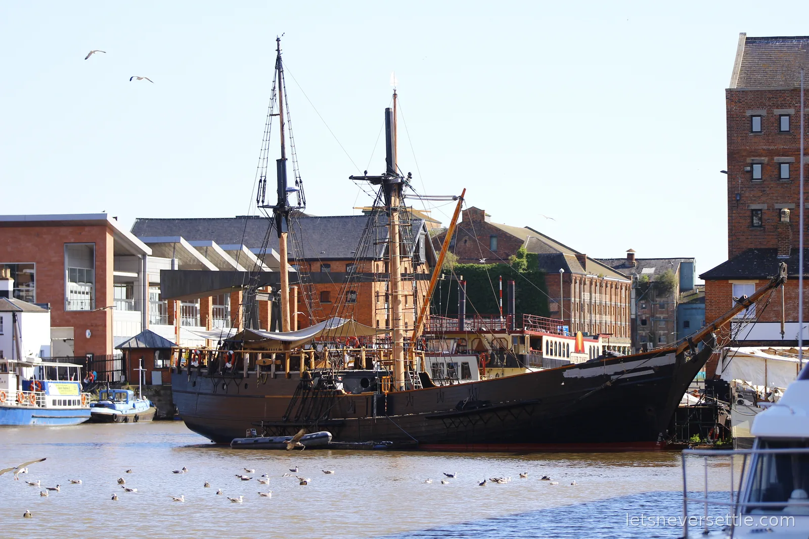 A dark-hulled two-masted tall ship moored at Gloucester Docks, red-brick Victorian warehouses behind and gulls on the water
