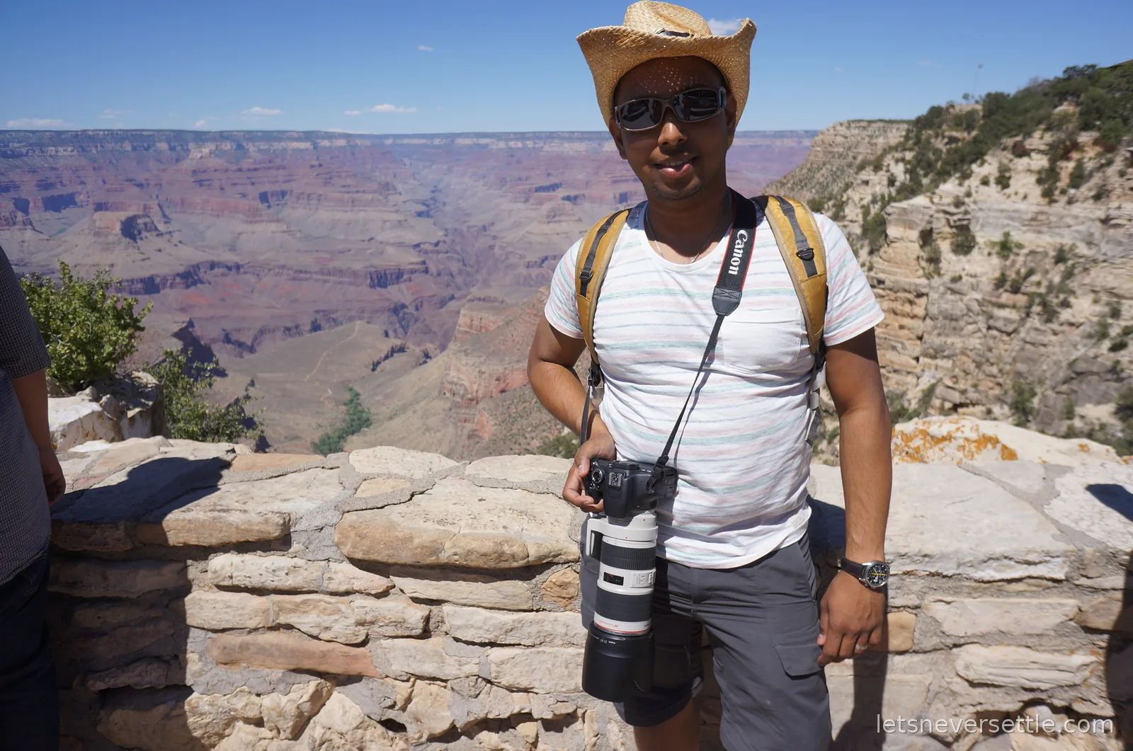 Dileep in a straw hat standing at the stone rim wall with a camera, canyon buttes behind