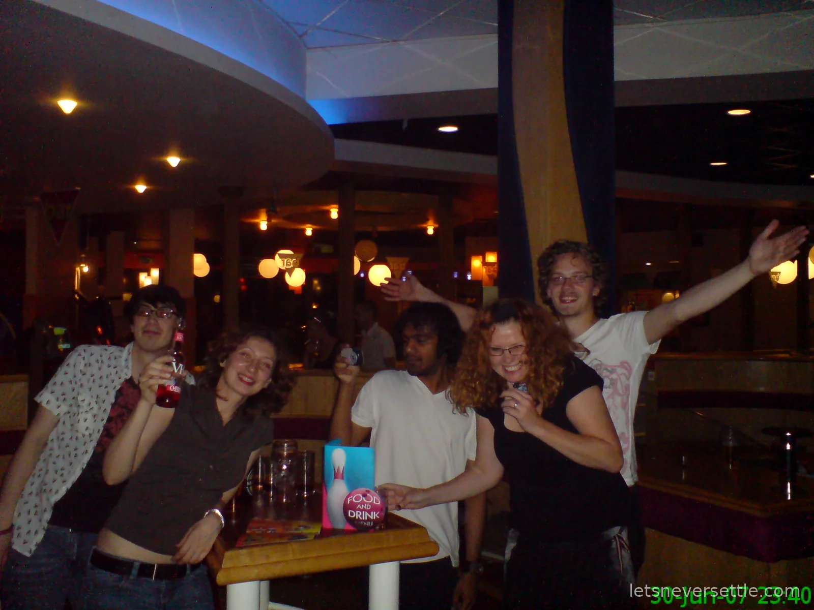 Five friends posing and cheering around a small table of drinks in the bar of a dimly lit bowling alley, a food and drink card standing on the table
