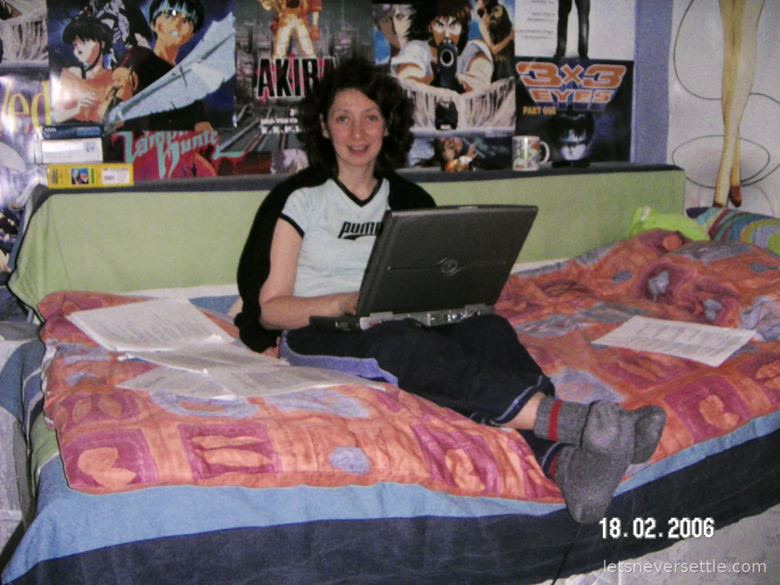 Inga sitting on a bed with a laptop on her knees, printed coursework pages spread around her and a wall of anime posters including Akira and 3x3 Eyes behind her