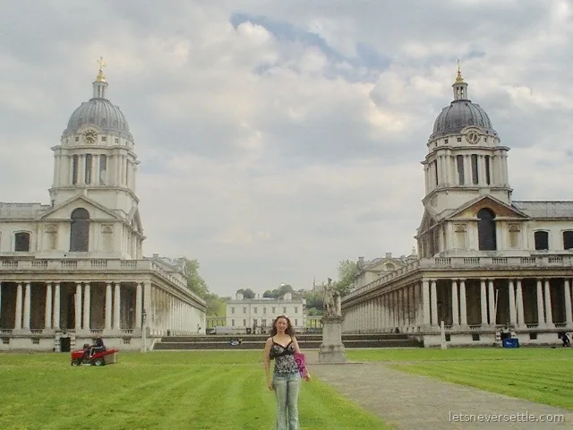 A woman standing on the wide green lawn between the two domed baroque blocks of the Old Royal Naval College in Greenwich, with the white Queen's House framed in the gap beyond