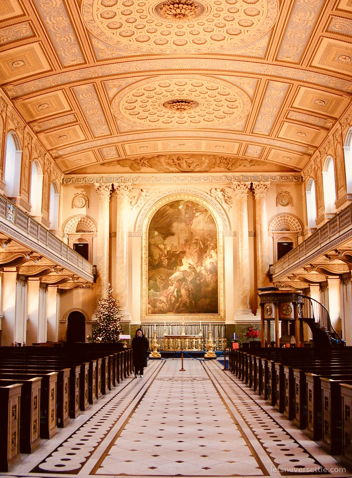 The long aisle of the Old Royal Naval College chapel in Greenwich, gilded coffered ceiling above, a tall altarpiece painting behind the altar and a lit Christmas tree to the left