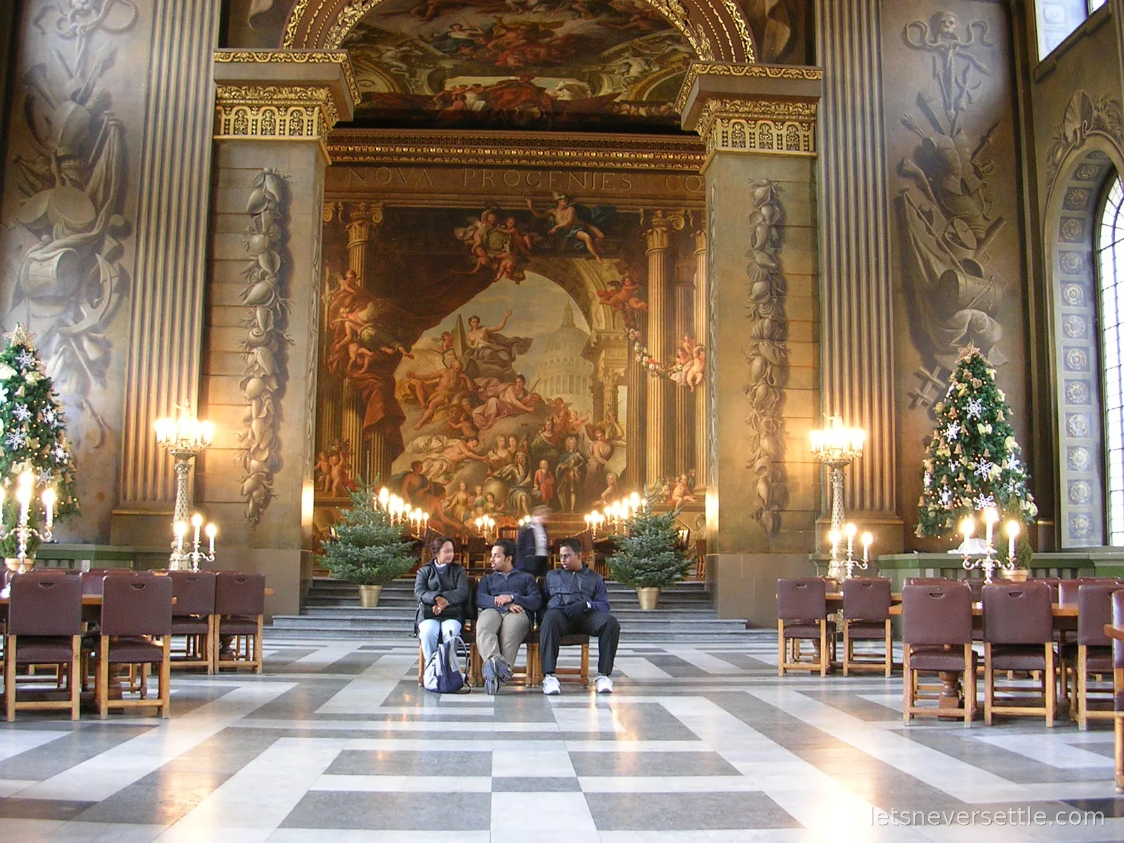 Three visitors sitting on chairs in the middle of the Painted Hall at Greenwich, the vast baroque wall painting and candelabra behind them and decorated Christmas trees on both sides