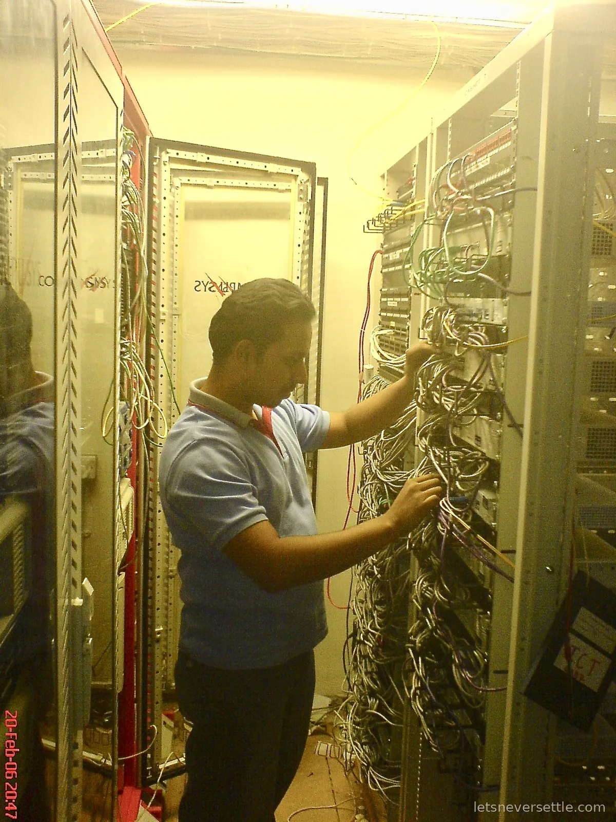 Dileep in a blue polo shirt and lanyard reaching into a dense bundle of patch cables on an open server rack in a machine room