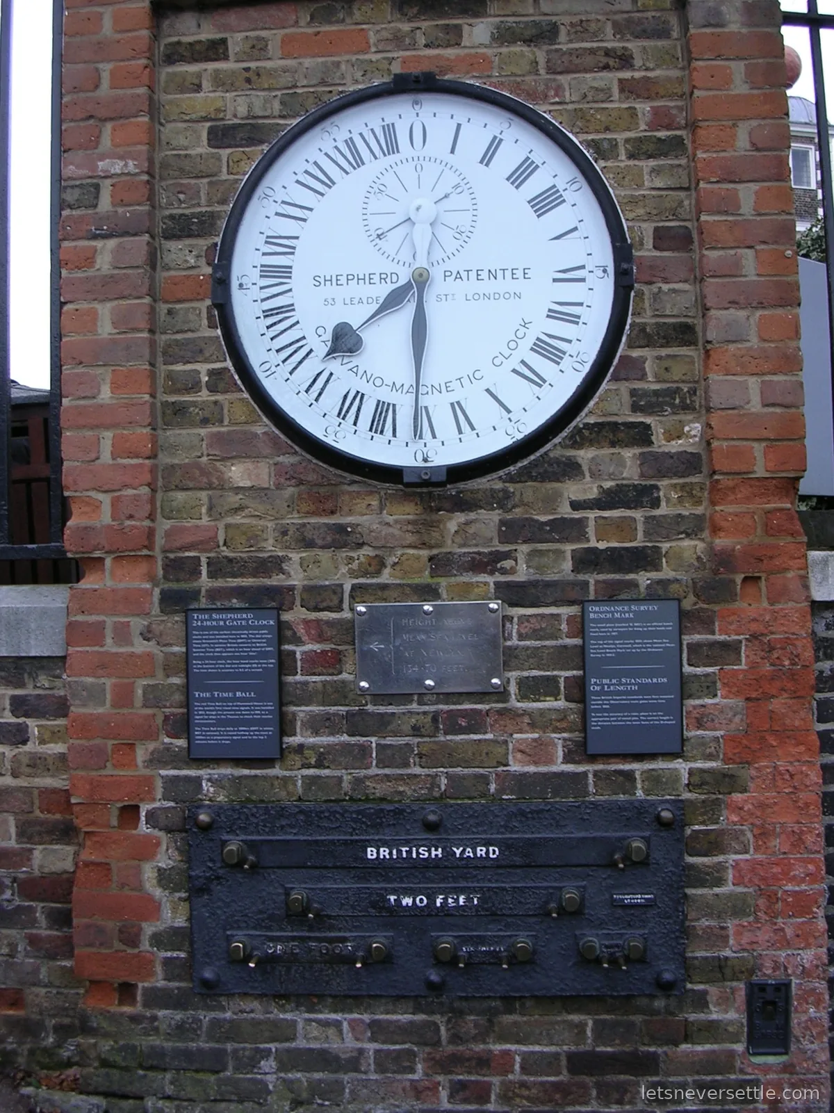 The Shepherd Gate 24 hour galvano-magnetic clock mounted on the brick wall at the Royal Observatory Greenwich, with the brass public standards of length for the British yard, two feet and one foot fixed below it