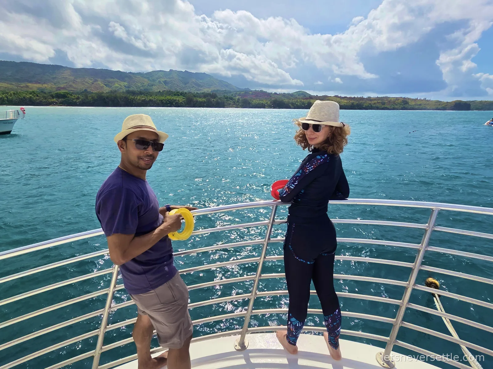 Dileep and Inga in sun hats on the white bow of a boat off Guam, holding yellow and red dive rings, green hills across the turquoise water