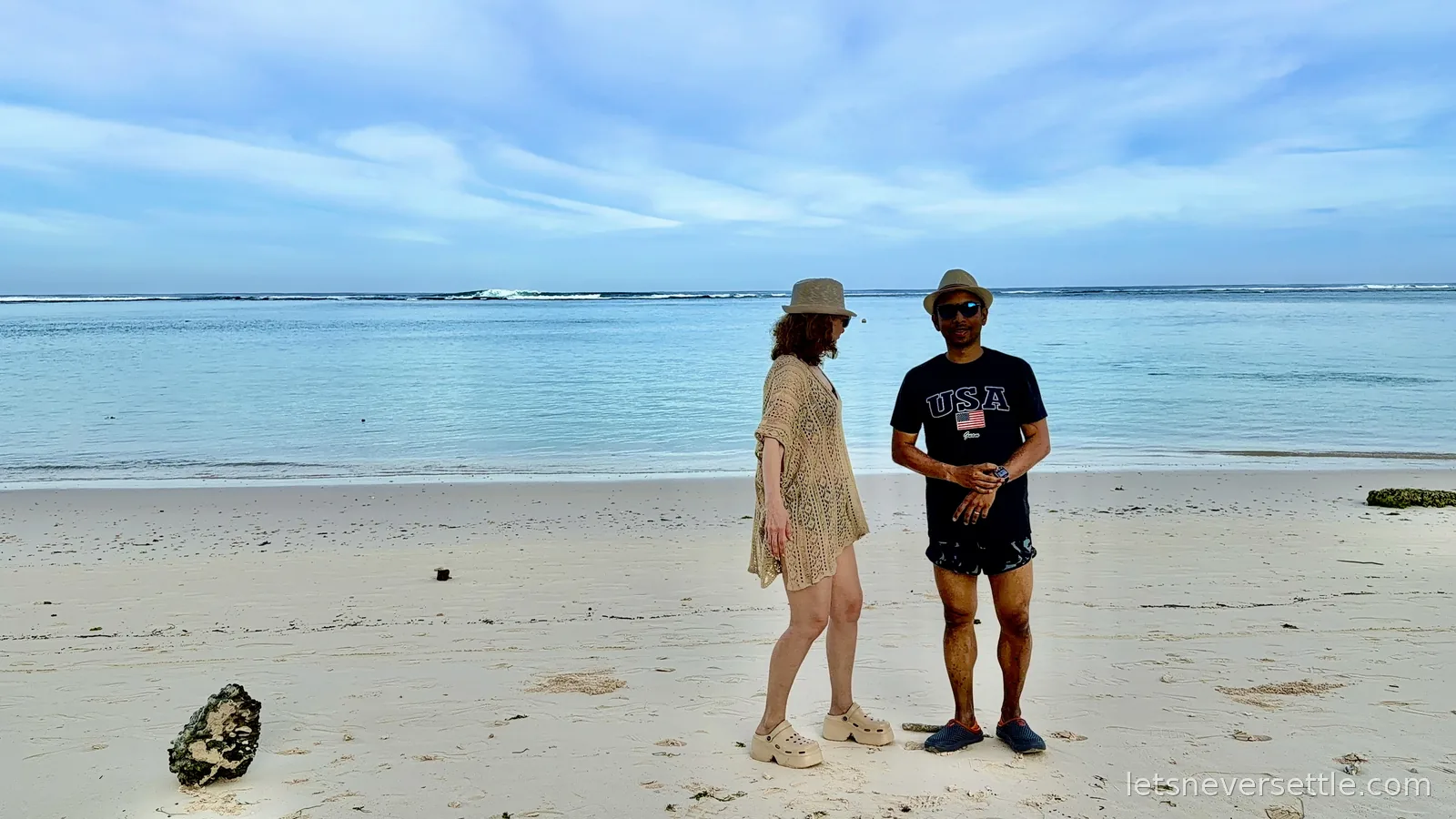 Inga in a crochet cover-up and Dileep in a USA t-shirt standing on empty white sand, the reef line breaking far out behind them