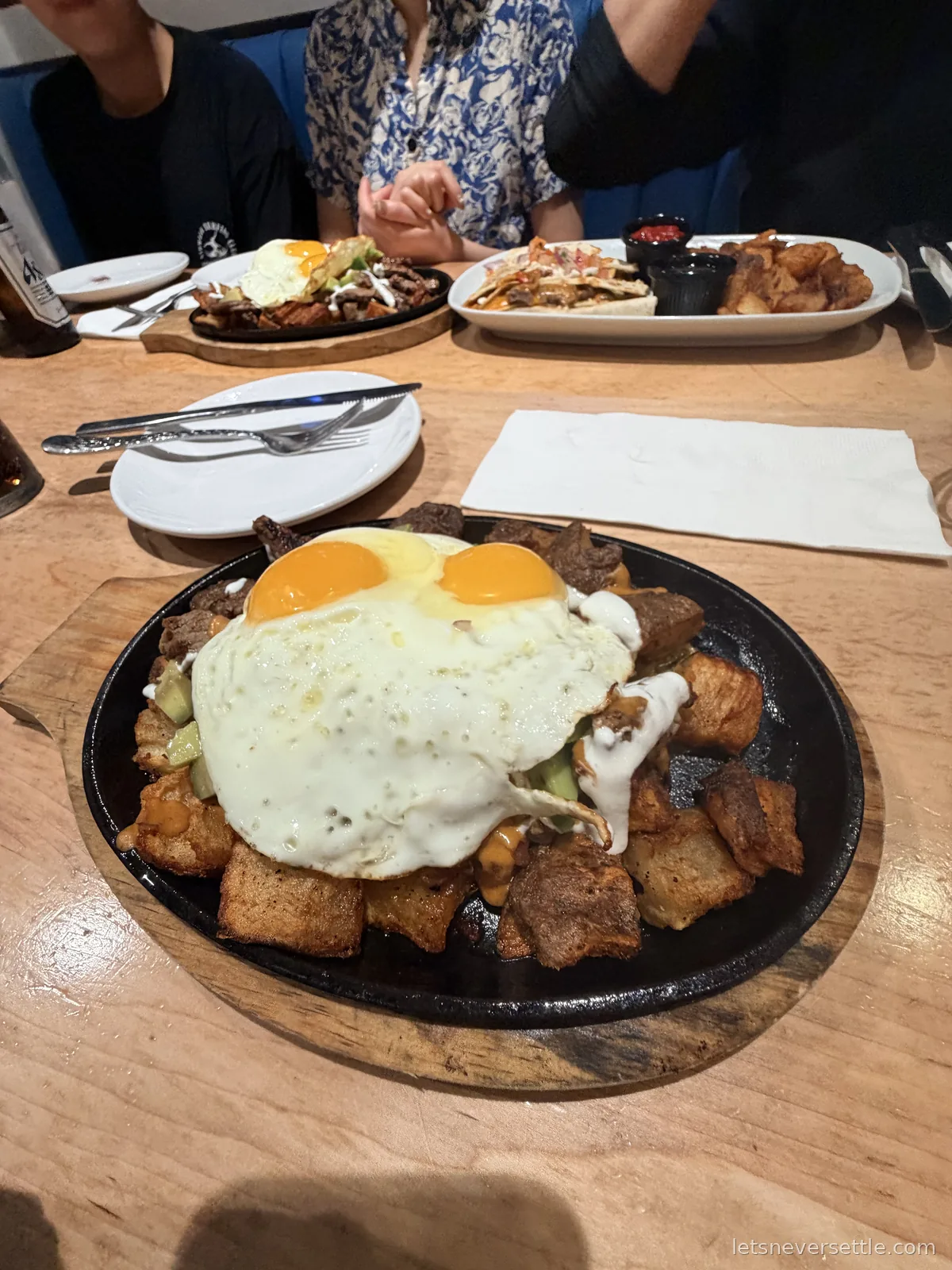 A cast iron skillet of fried potatoes and beef topped with two fried eggs, with loaded fries and another sizzling plate on the table behind