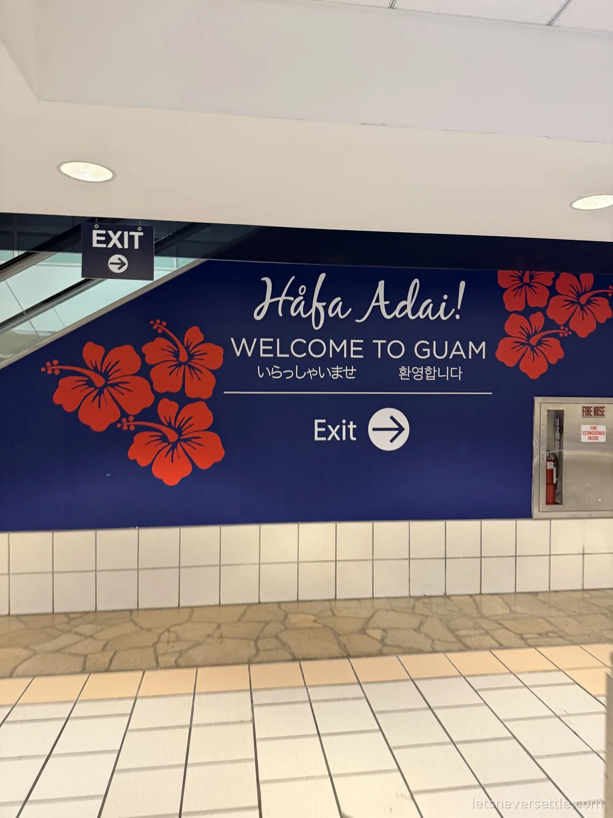 Blue Hafa Adai Welcome to Guam sign with red hibiscus flowers at the airport arrivals exit