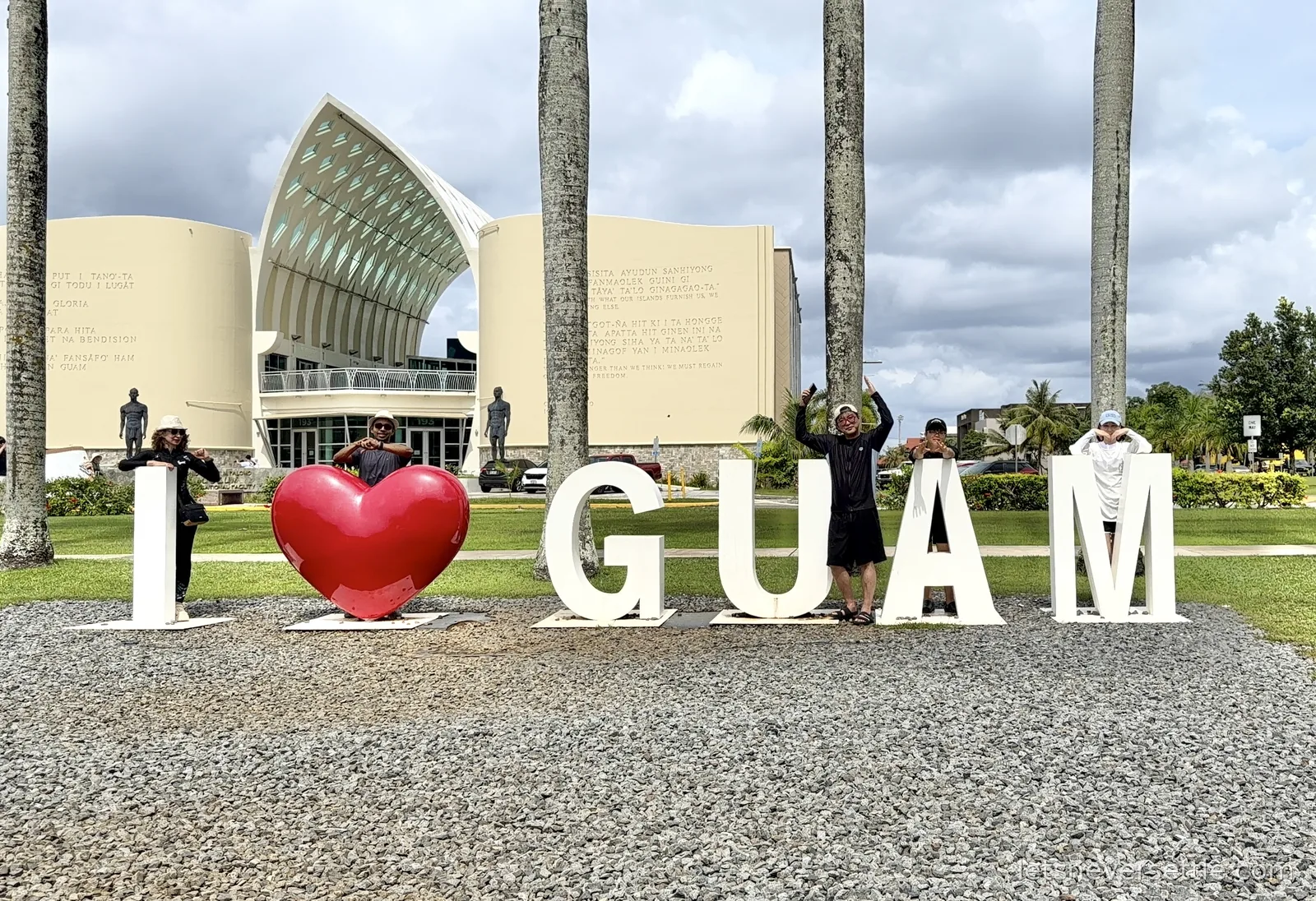 Large white I love Guam letters with a red heart on gravel in Hagatna, people posing between them, palm trees and a modern building with a curved sail-shaped roof behind