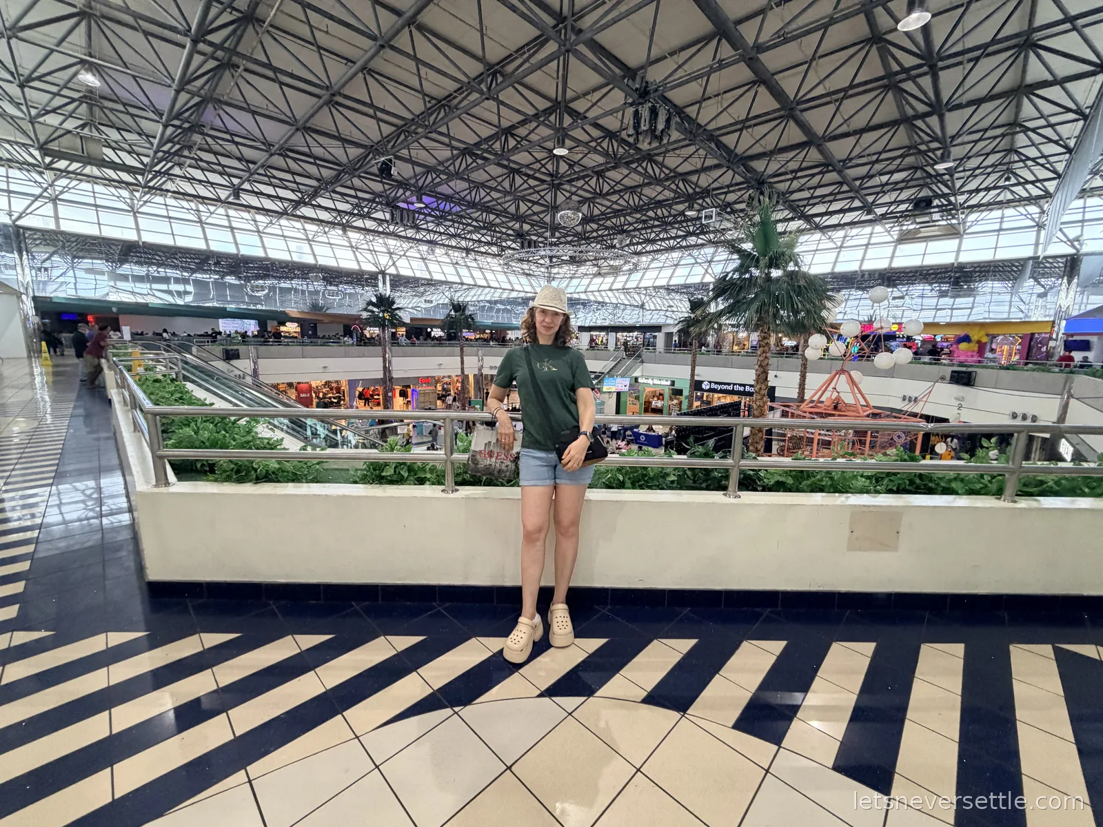 Inga standing on the upper level of Micronesia Mall in Guam, indoor palm trees and shops under a steel-truss glass roof