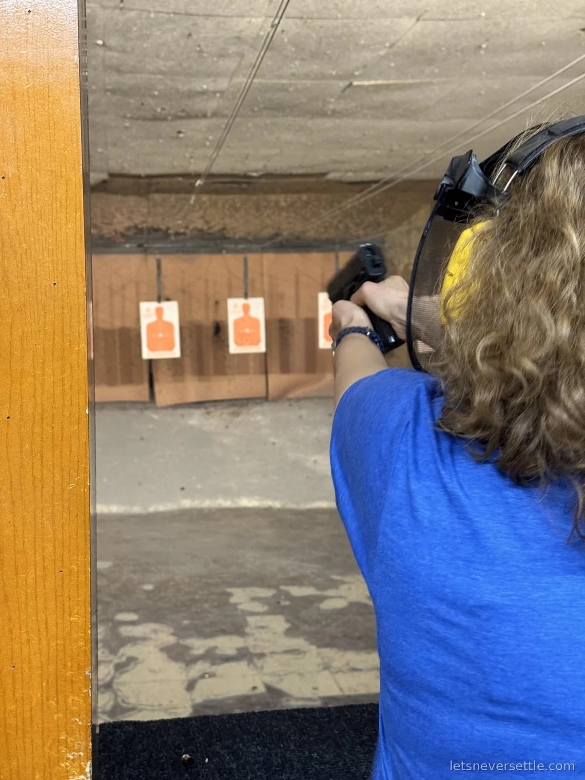 Inga from behind in ear defenders and safety glasses, aiming a pistol two-handed down an indoor range lane at three paper targets
