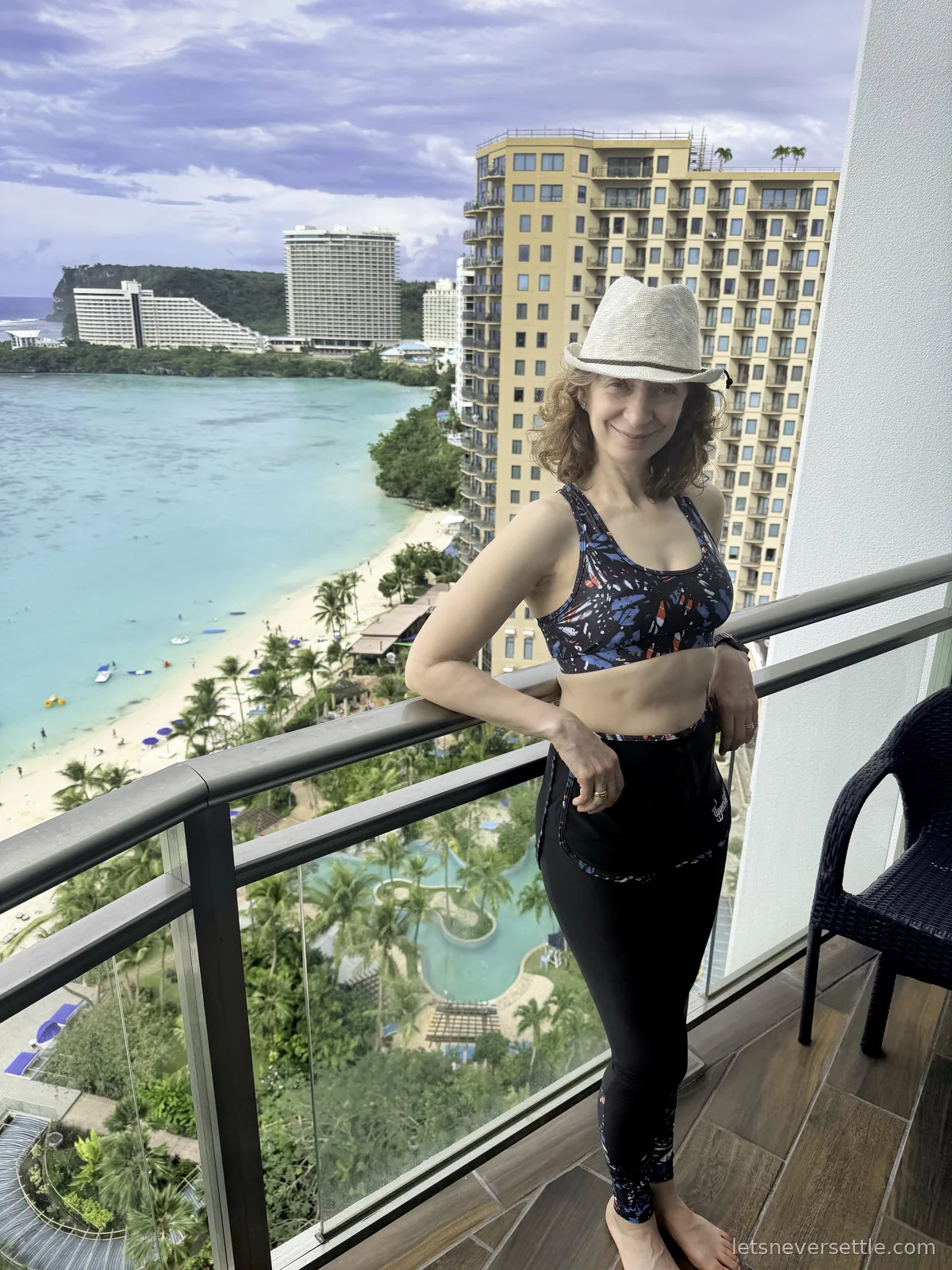 Inga in a straw hat on a high hotel balcony above Tumon Bay, the turquoise lagoon, palm-fringed beach and resort pool laid out below