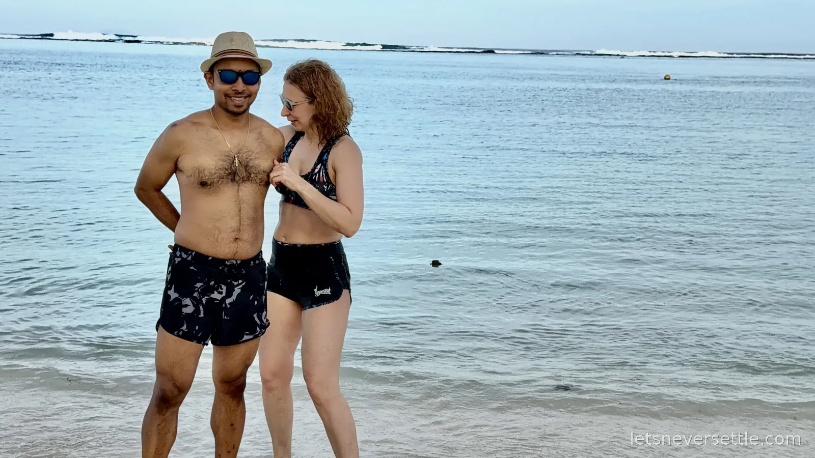 Inga and Dileep in swimwear standing in calm shallow water at Tumon Bay, waves breaking far out on the reef