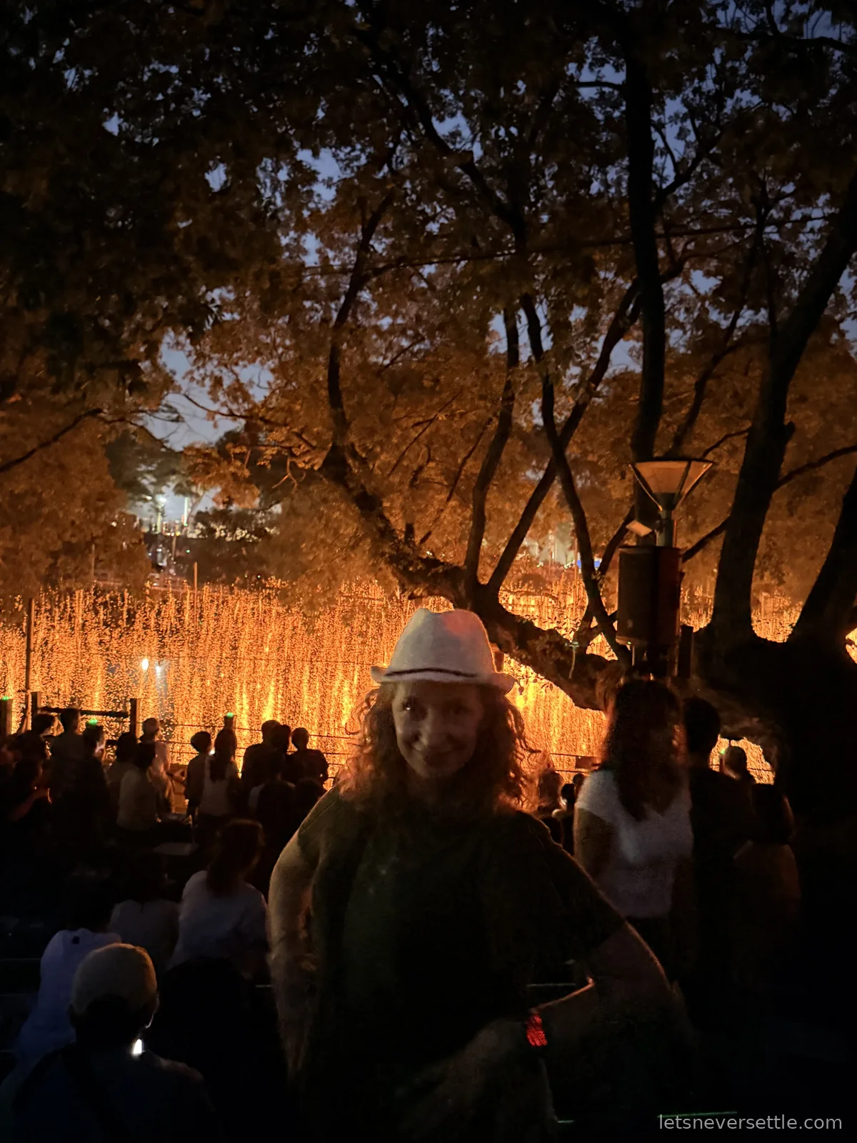 Inga in a white hat standing in front of a glowing curtain of orange embers falling from ropes over the pond at night in Haman