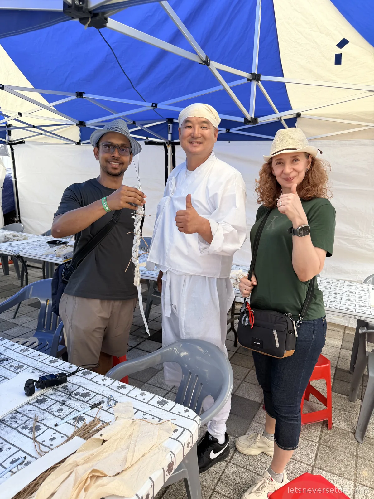 Inga and Dileep giving thumbs up with a smiling stallholder dressed in traditional white at a festival tent in Haman