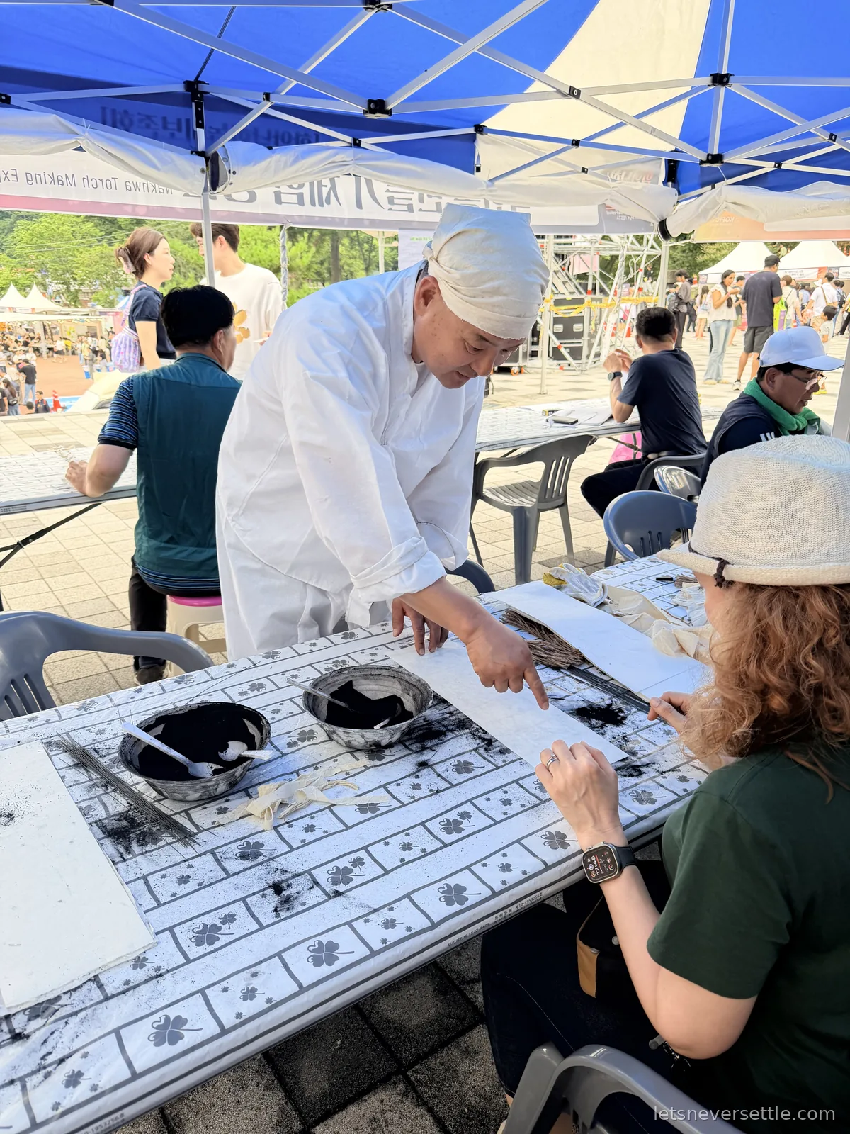 Inga at a festival workshop table in Haman packing charcoal powder into paper strips while a man in traditional white robes leans over and points
