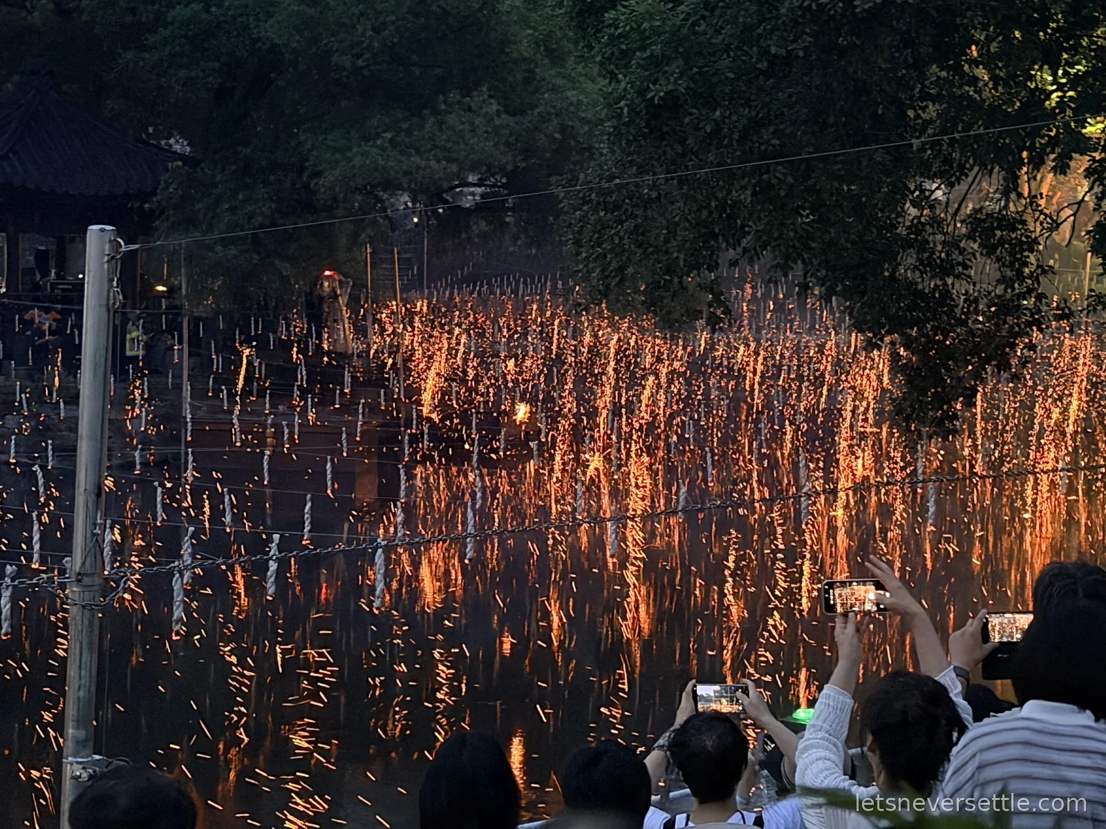 Ropes strung over a pond at dusk in Haman shedding thousands of orange embers into the water, crowds filming below