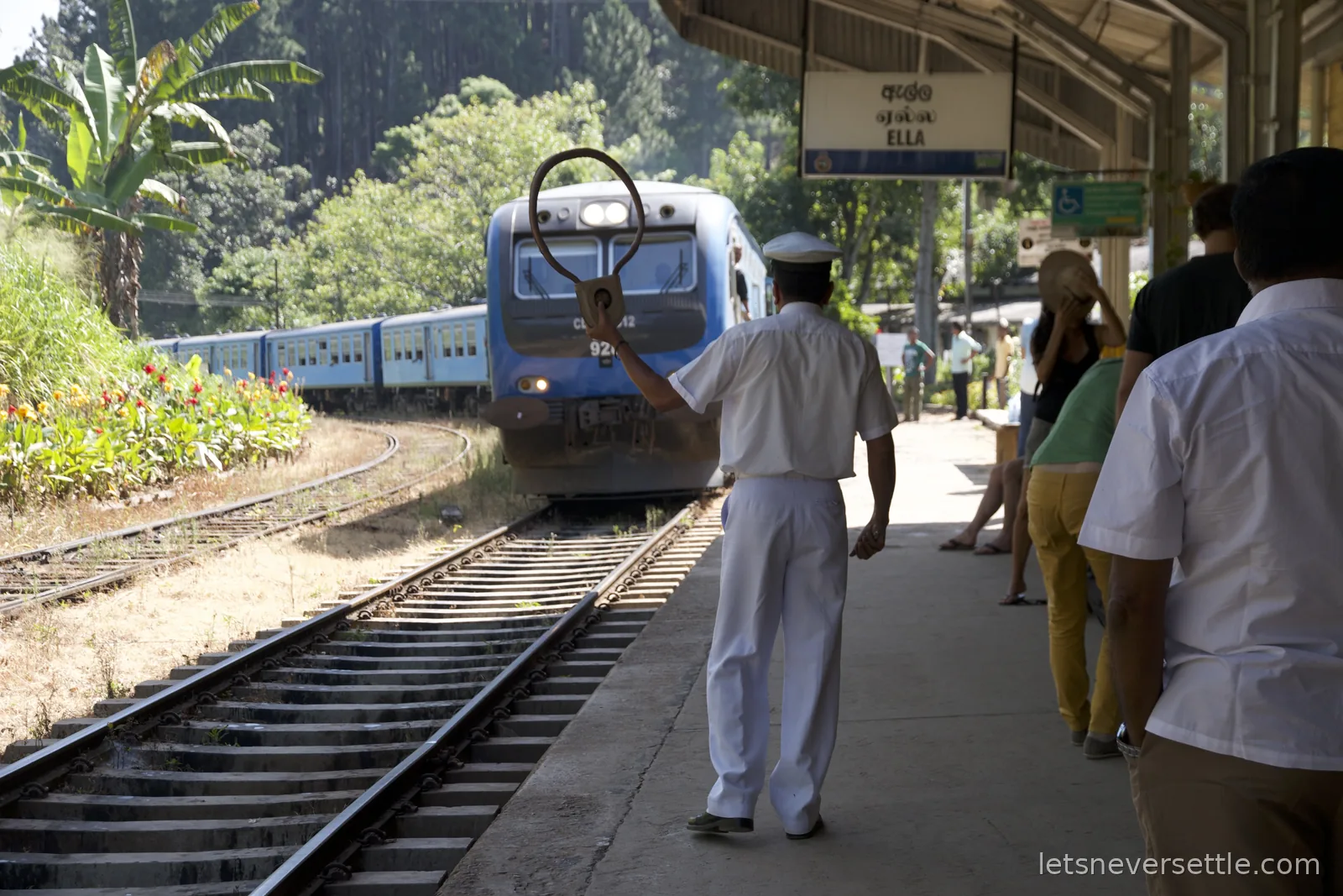 A stationmaster in white uniform holding up a hooped token as a blue train pulls into Ella station