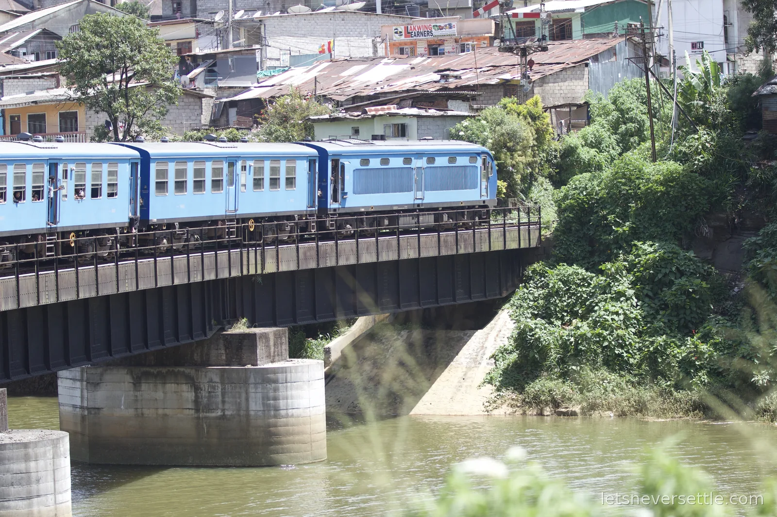 Blue train crossing a black girder bridge over a river with a hillside town stacked up behind