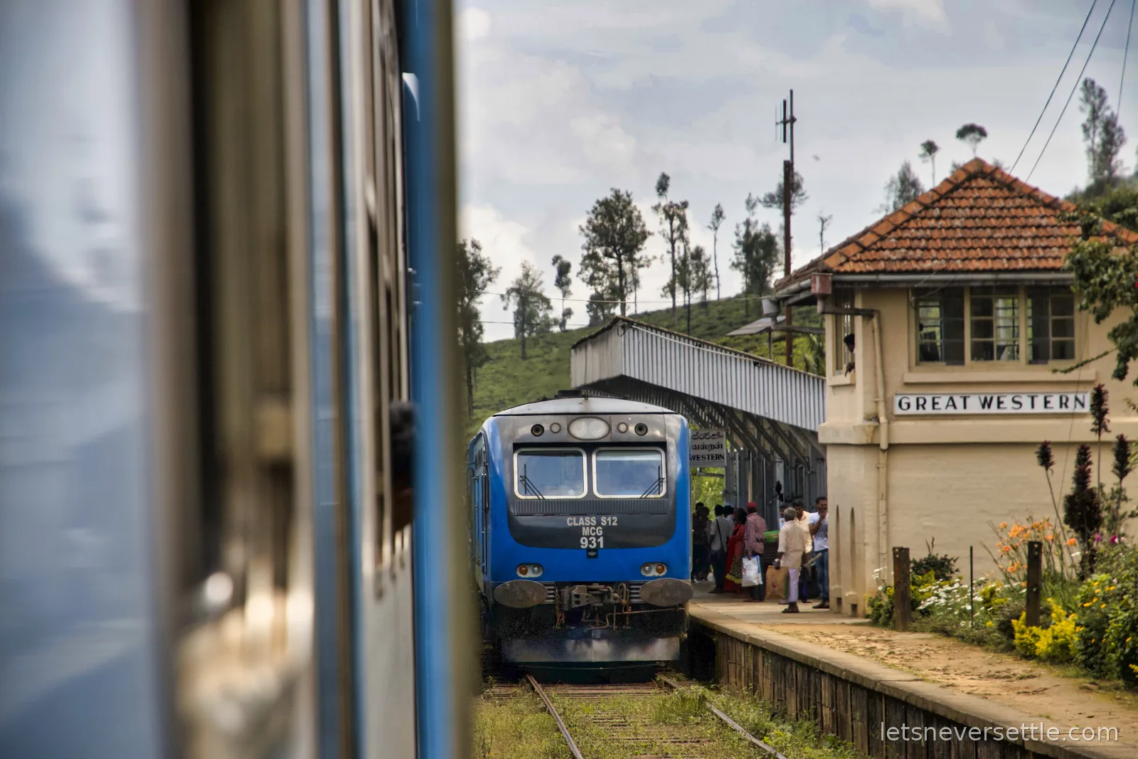 A blue train waiting beside the old station house at Great Western, seen past the carriages of another train