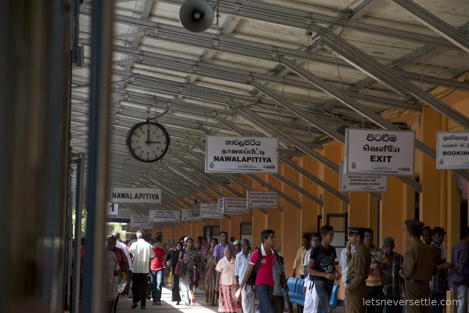 Nawalapitiya station platform with hanging trilingual signs, a clock and crowds of passengers under the iron roof