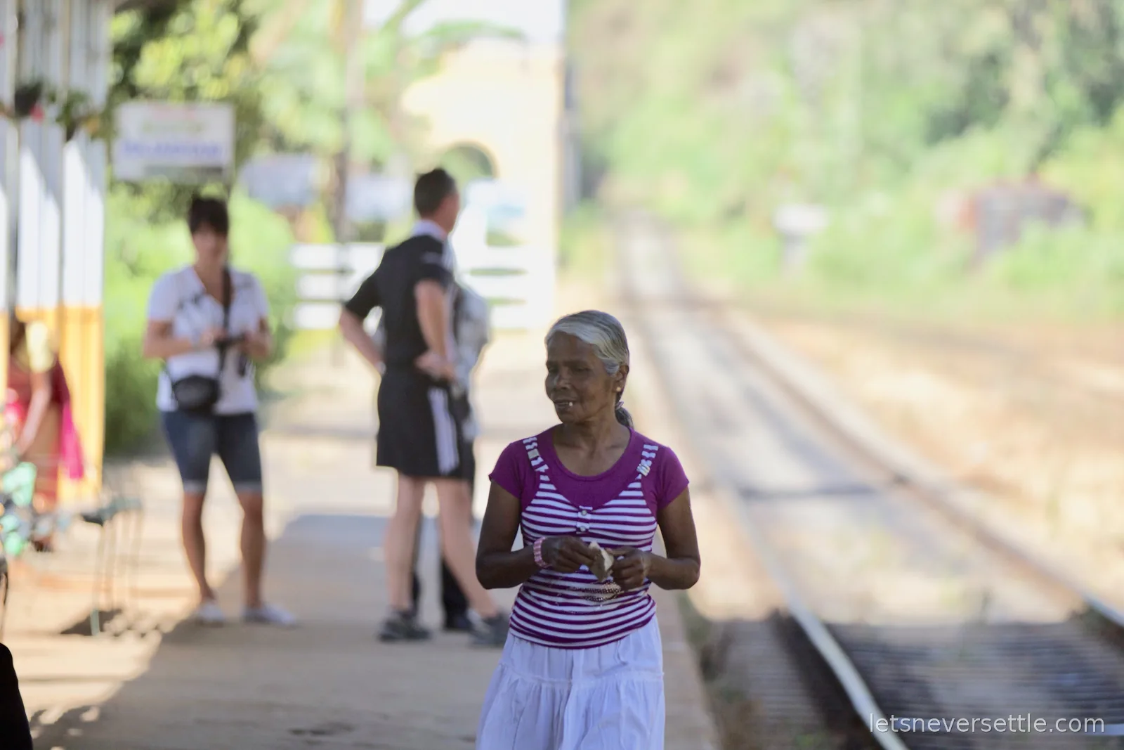 A woman in a striped pink top walking along a station platform, other travellers and the line soft in the background