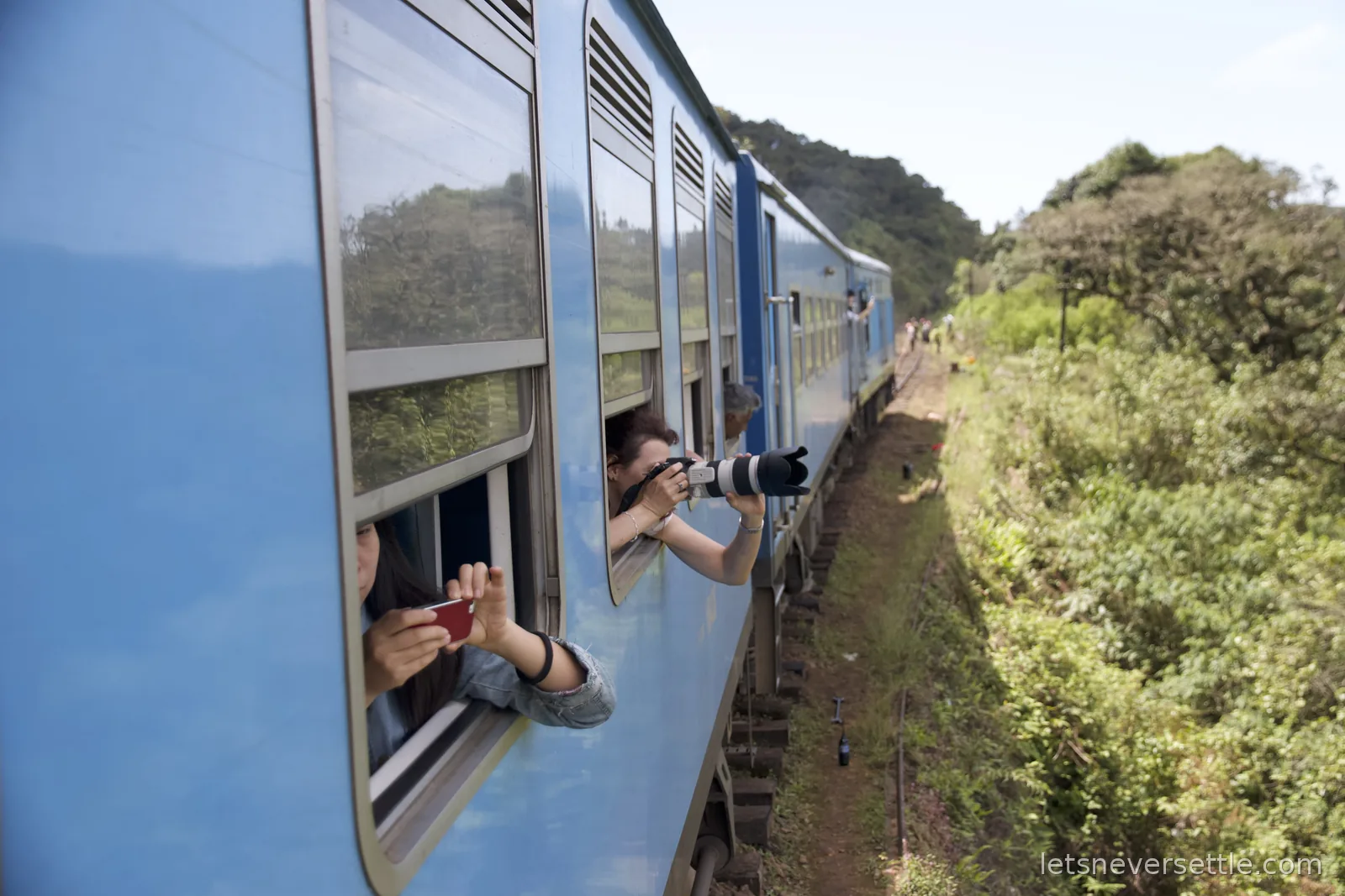 Passengers leaning out of the blue train windows with a camera and a phone pointed down the line