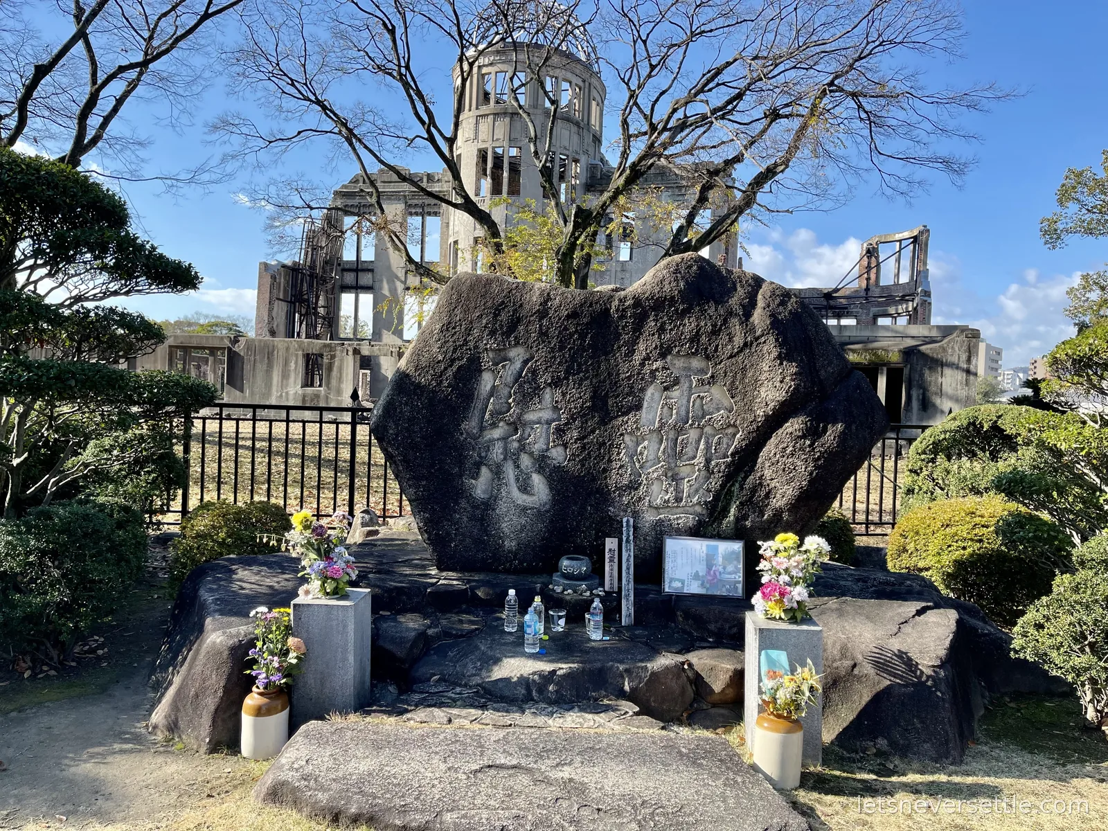 A stone memorial with fresh flowers and offered bottles of water standing in front of the ruined A-Bomb Dome