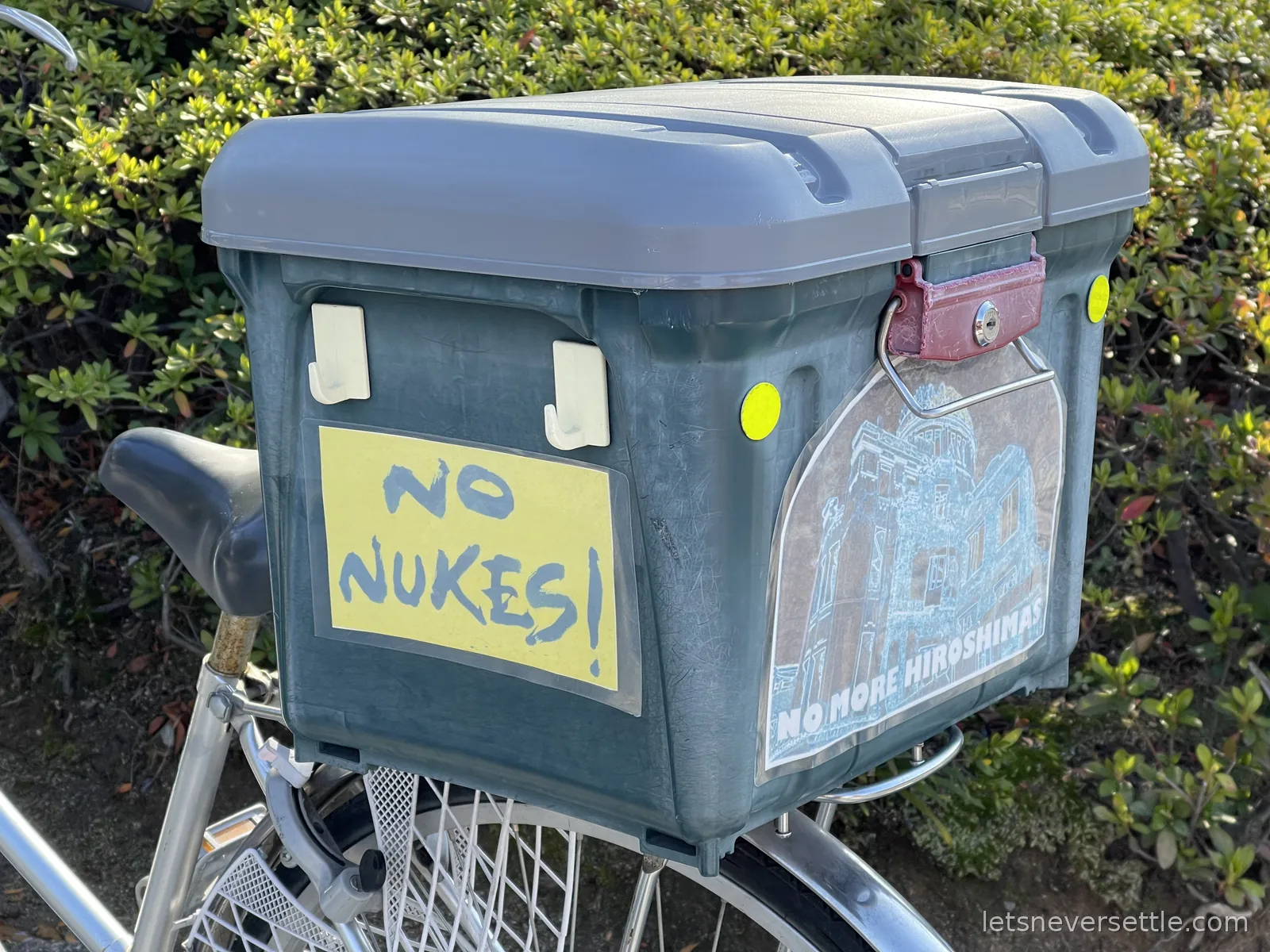 A hand-painted No Nukes sign taped to a delivery box on a parked bicycle, with No More Hiroshimas printed on its side