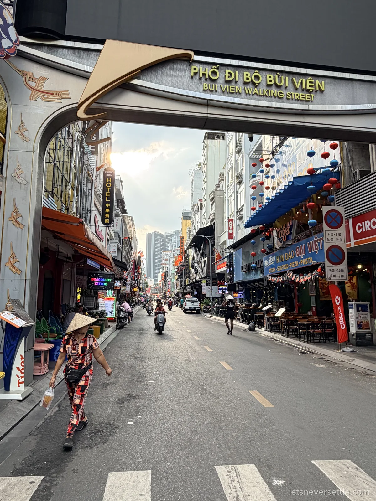 The gold Bui Vien Walking Street archway spanning an almost empty District 1 street, a woman in a conical hat crossing beneath it and red and blue lanterns strung above the bars on the right