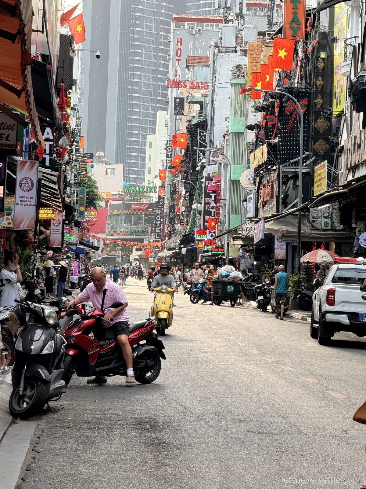 Looking down Bui Vien street past parked scooters and stacked hotel signs, Vietnamese flags hanging from the balconies and a man resting on a red motorbike in the foreground