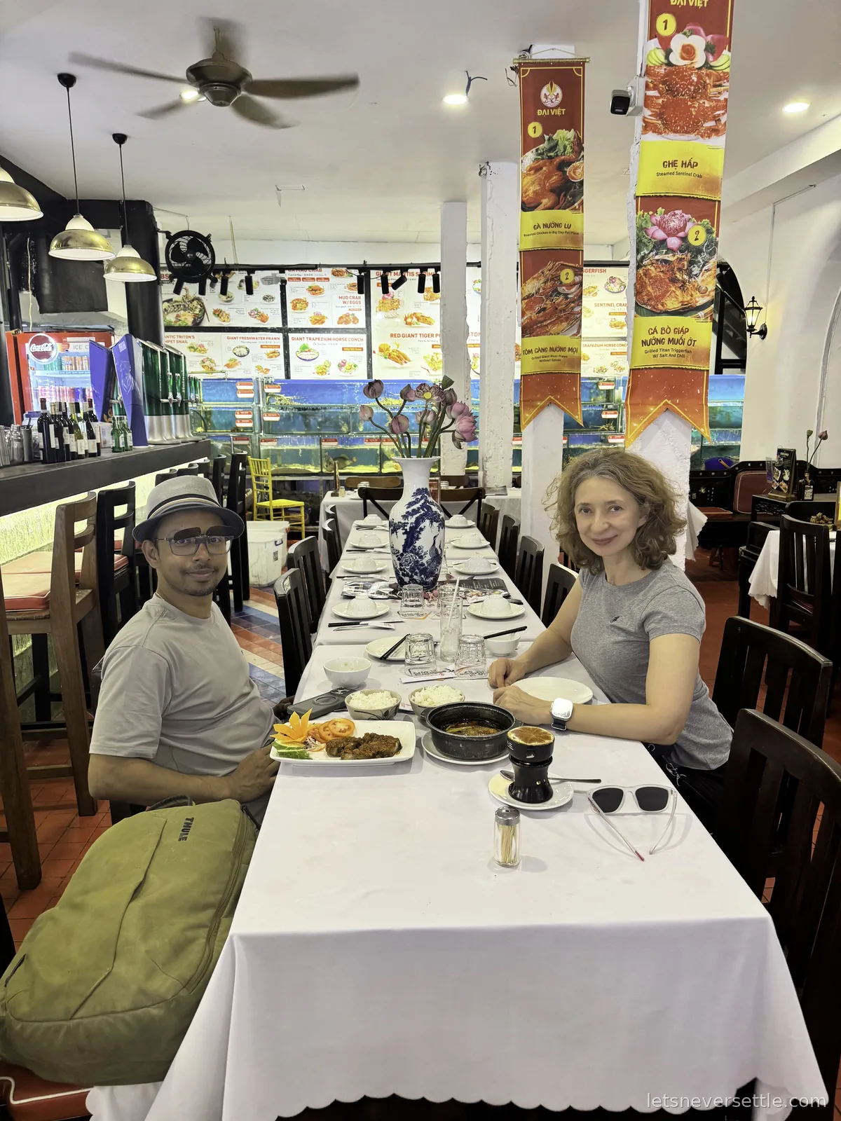 Dileep in a bucket hat and Inga seated at opposite sides of a long white banquet table in a Vietnamese restaurant, a black clay pot and rice in front of them and fish tanks along the back wall
