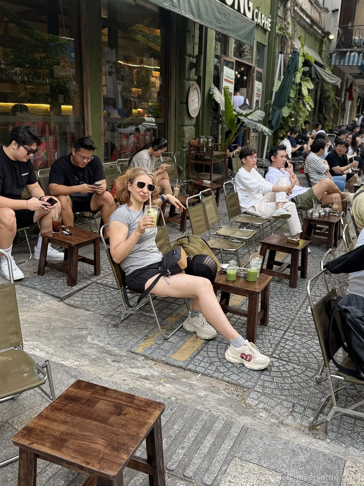 Inga sitting on a low canvas chair on the pavement outside a green-fronted ca phe holding an iced green drink, surrounded by young locals on stools with their own iced coffees